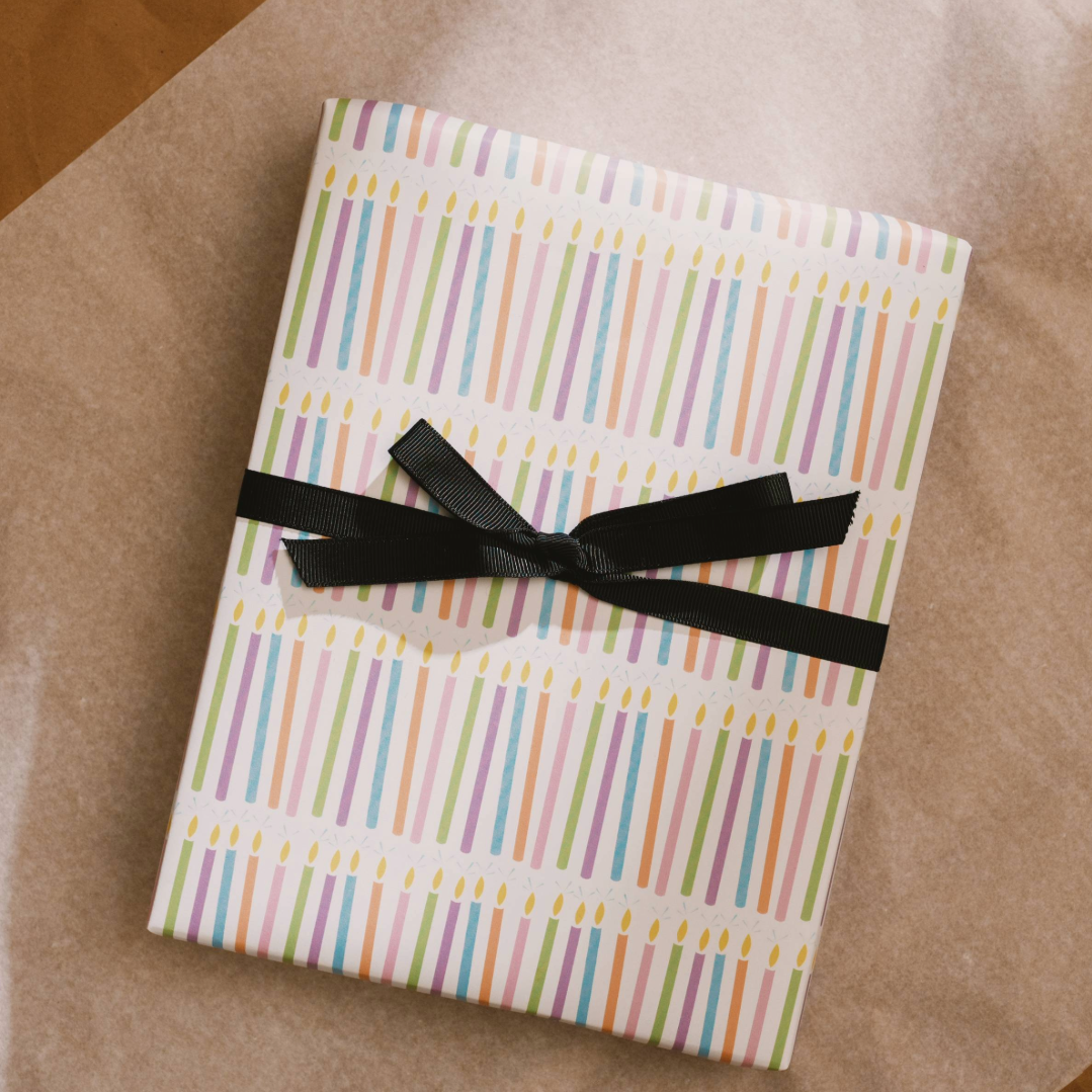 A gift wrapped with illustrated pastel birthday candles wrapping paper. A black ribbon is tied in a bow across the middle.