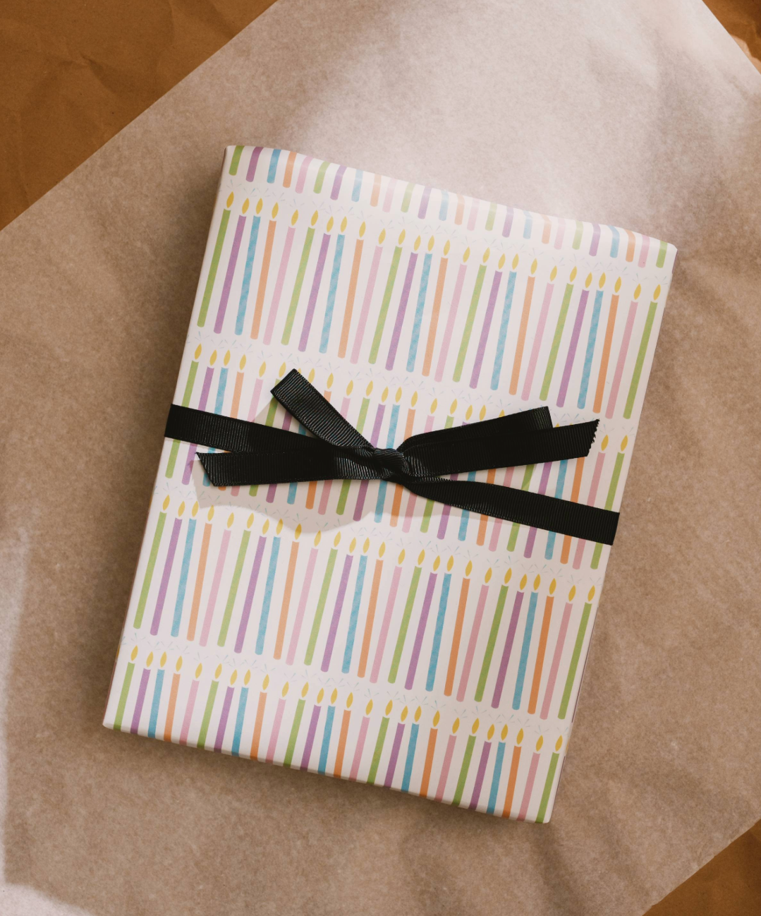 A gift wrapped with illustrated pastel birthday candles wrapping paper. A black ribbon is tied in a bow across the middle.
