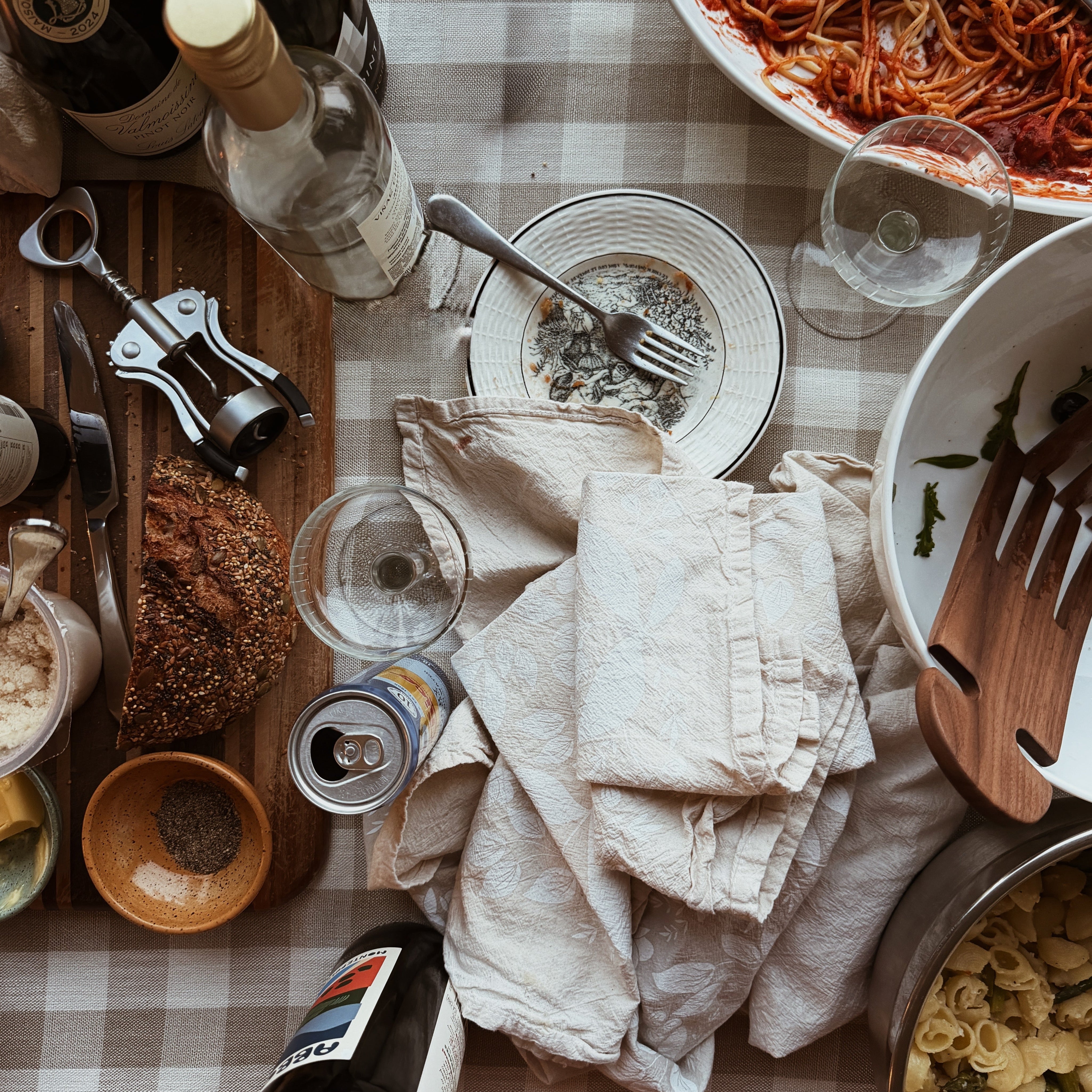 A cluttered dinner table with open wine bottles, empty plates, and serving dishes pushed to the edge of the table with a checkered tablecloth.