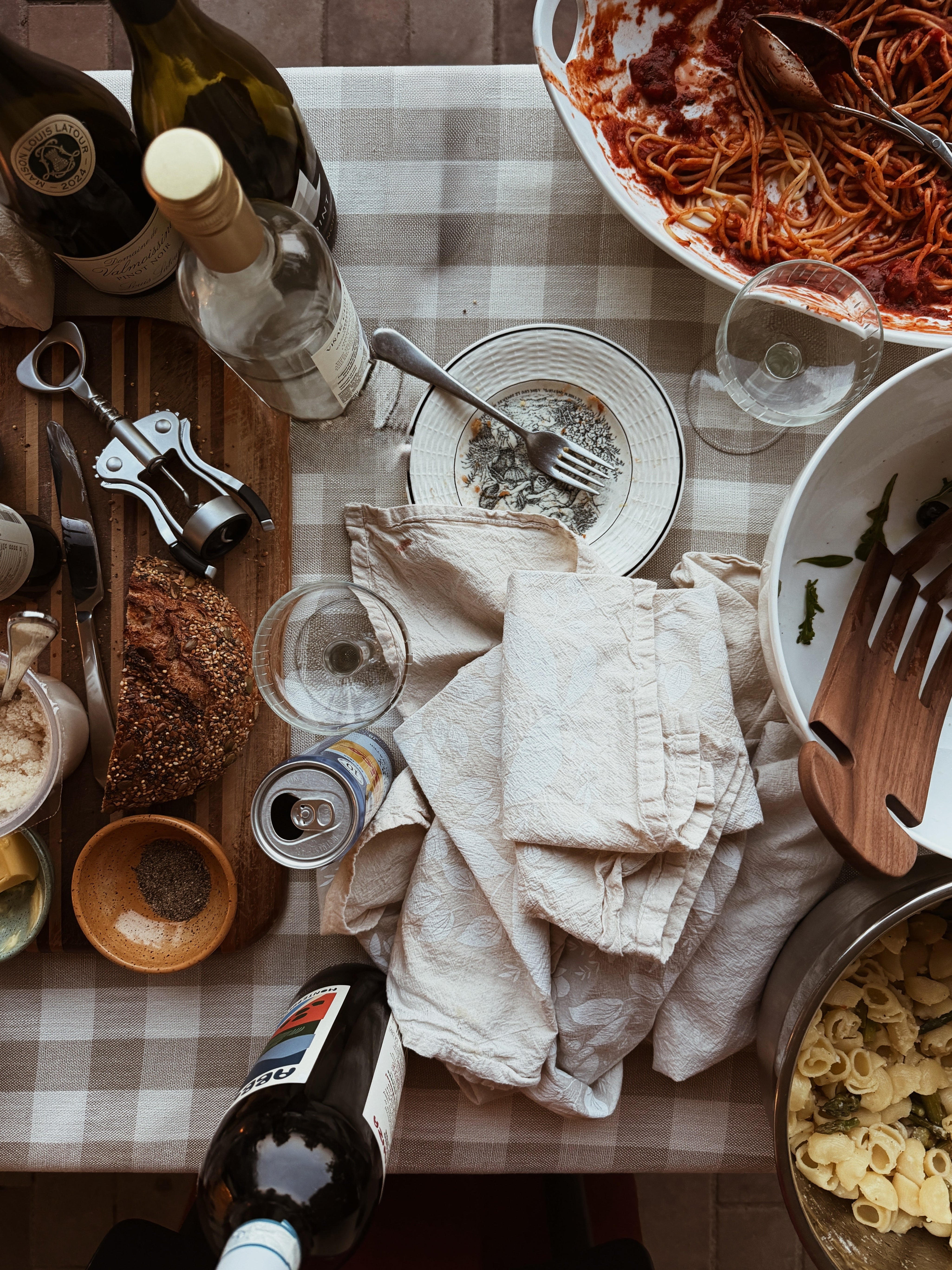 A cluttered dinner table with open wine bottles, empty plates, and serving dishes pushed to the edge of the table with a checkered tablecloth.
