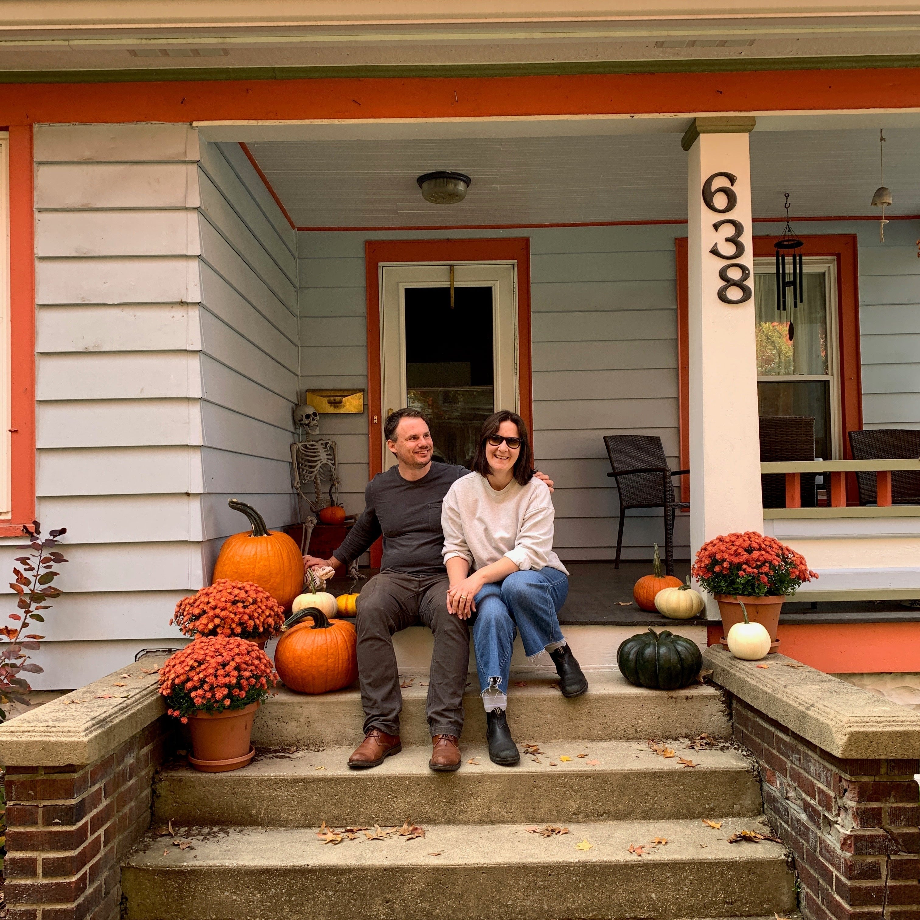 Susan and her husband Charlie sit on the porch of their old house, a light blue colonial-style house with reddish orange trim. There are pumpkins and potted chrysanthemums around them on the porch.