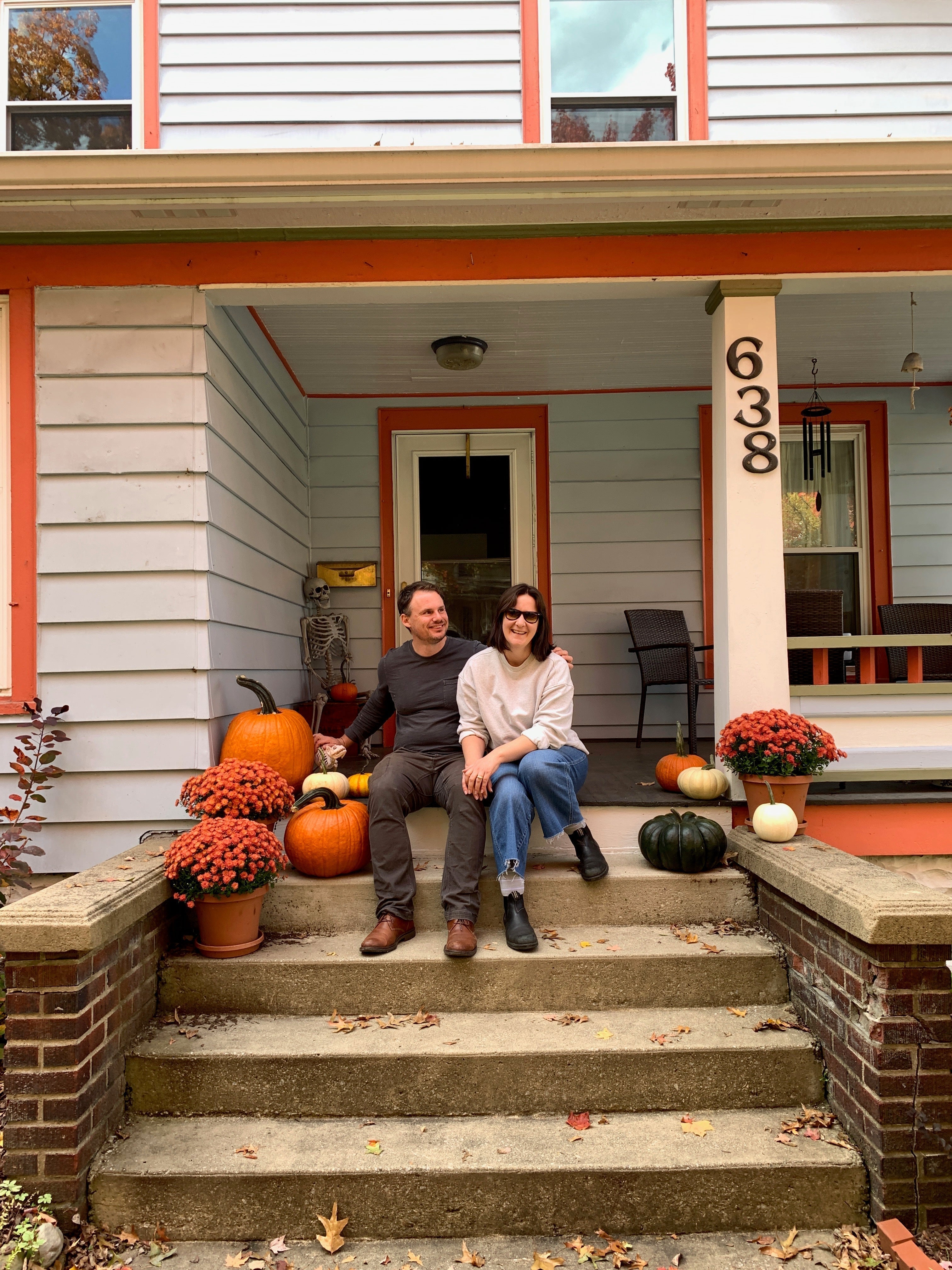 Susan and her husband Charlie sit on the porch of their old house, a light blue colonial-style house with reddish orange trim. There are pumpkins and potted chrysanthemums around them on the porch.
