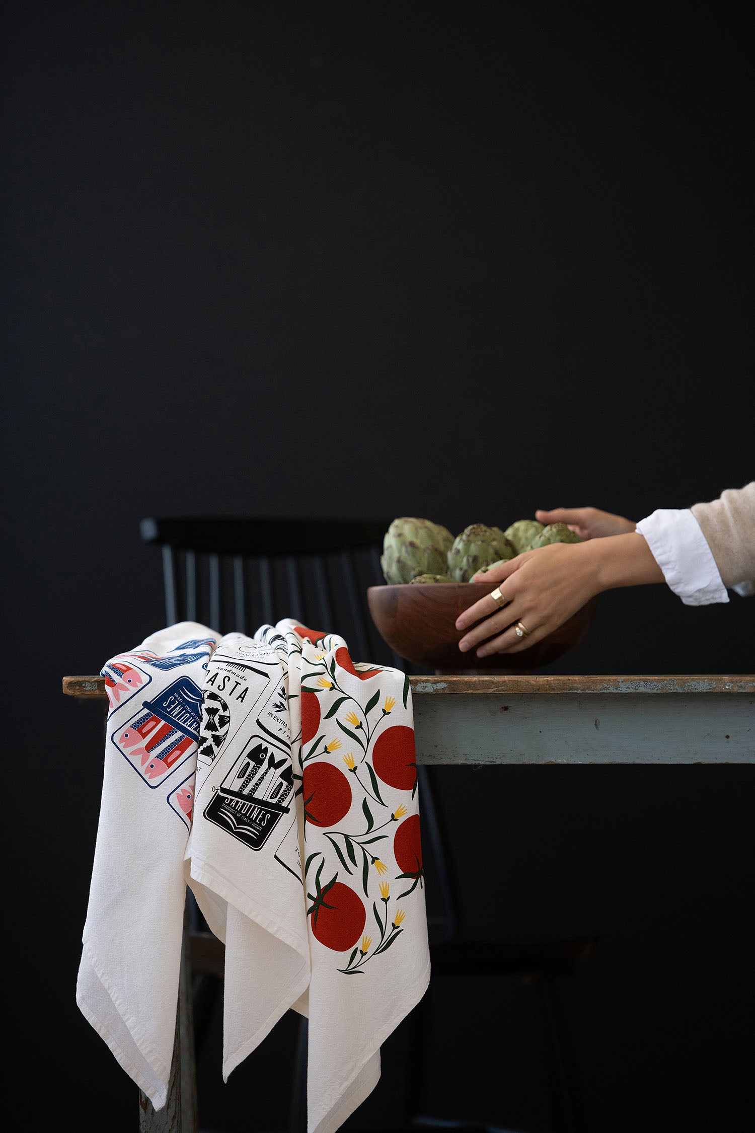 Hand arranging a bowl of artichokes on a table beside hand-illustrated cotton tea towels