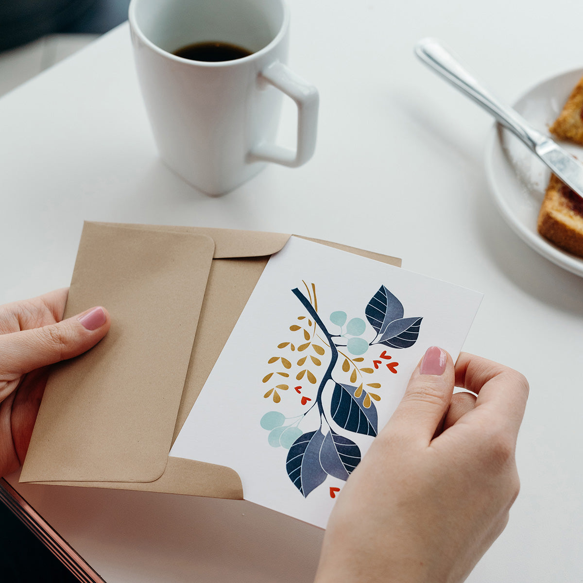 Hands holding a Hazelmade illustrated card and envelope beside a cup of coffee