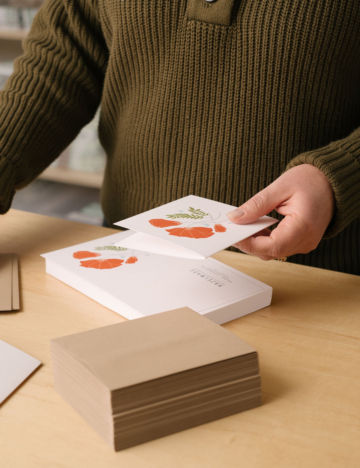 Hand-illustrated boxed greeting cards with orange floral design being packaged by hand