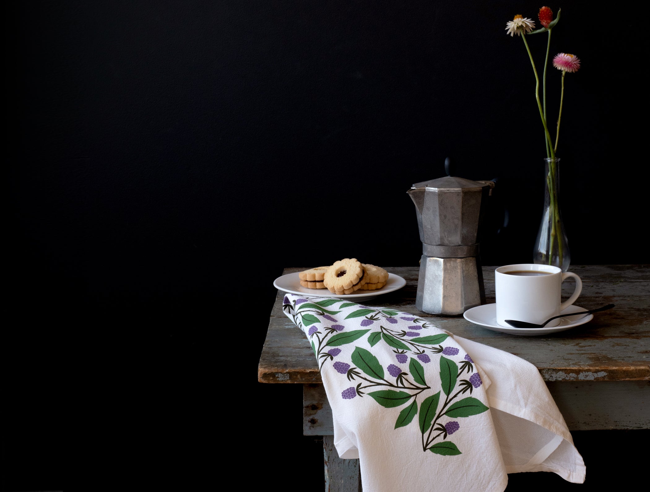 USA-made hand-illustrated cotton tea towel with berry print on a wooden table beside coffee and cookies