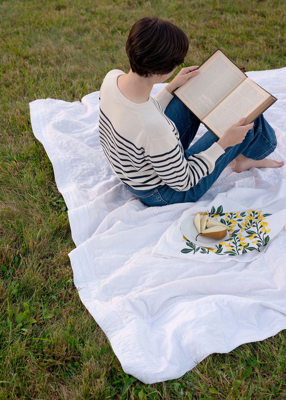Person reading on a white blanket outdoors beside a hand-illustrated tea towel and sliced pear