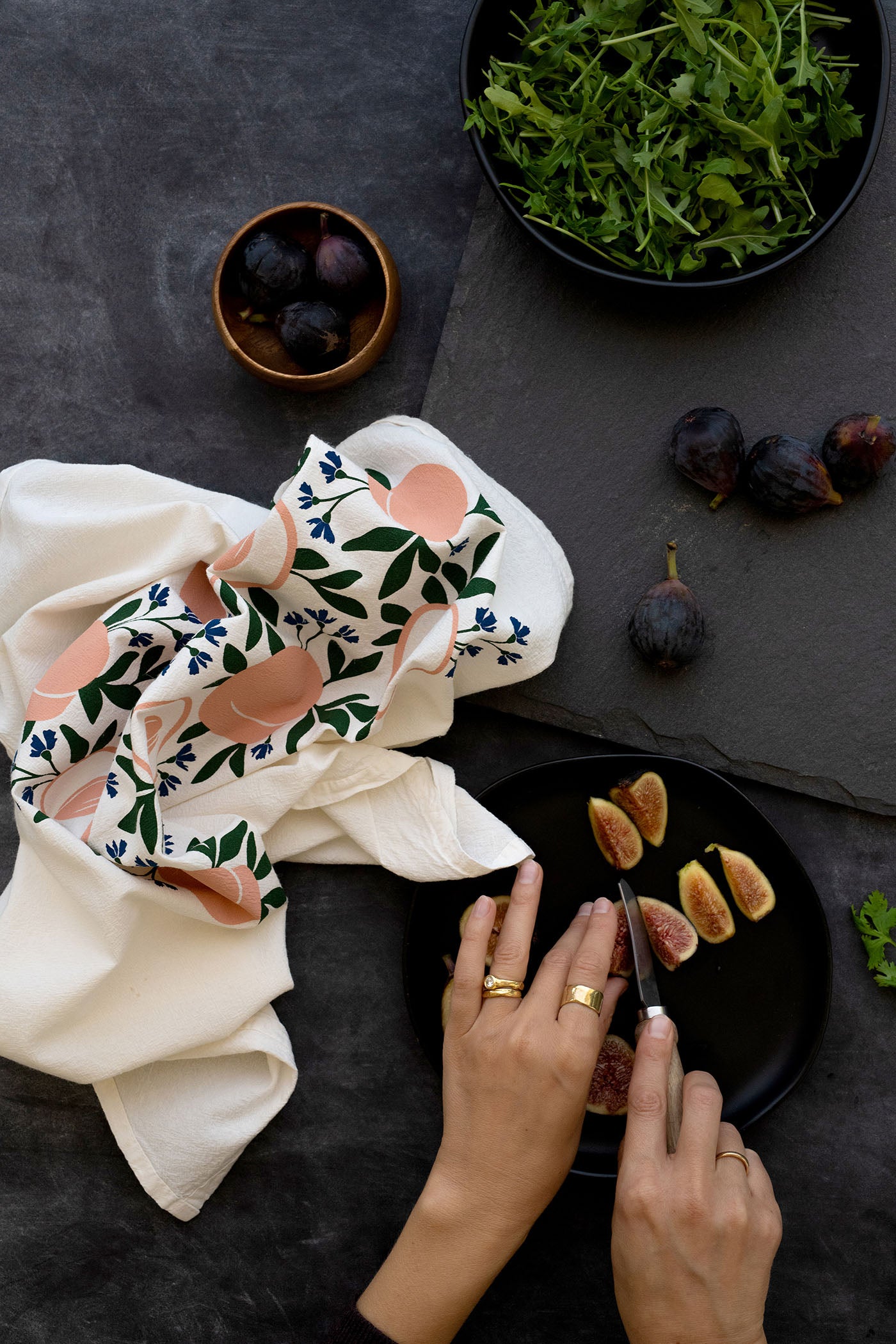Hazelmade peach-patterned tea towel styled with fresh figs and greens while a person slices figs on a dark tabletop