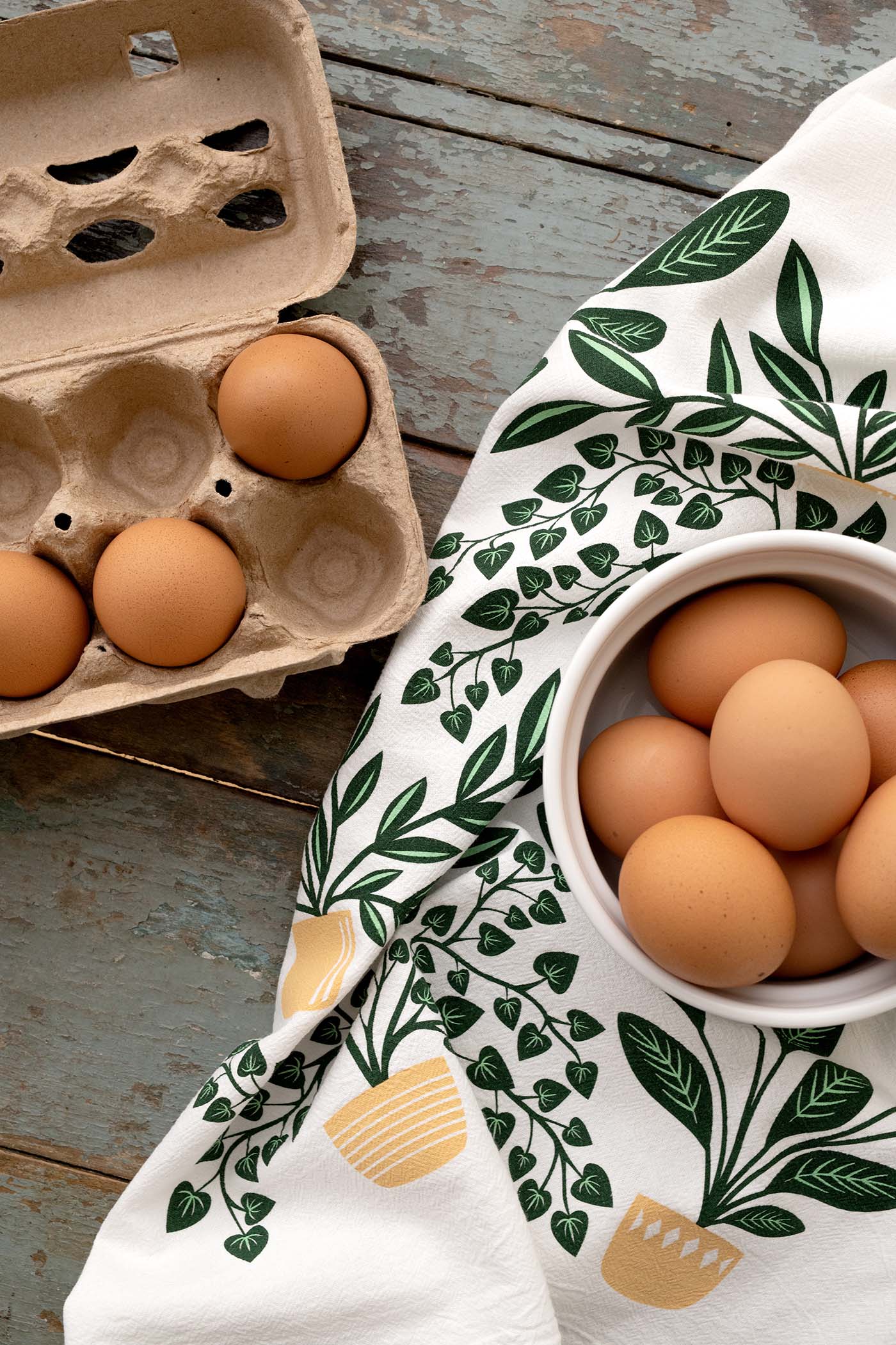 Hazelmade botanical tea towel styled with eggs in a carton and bowl on a rustic wooden surface