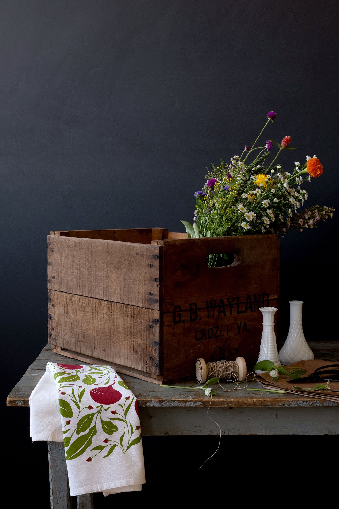 Hand-illustrated tea towel draped over a wooden table beside a flower-filled crate