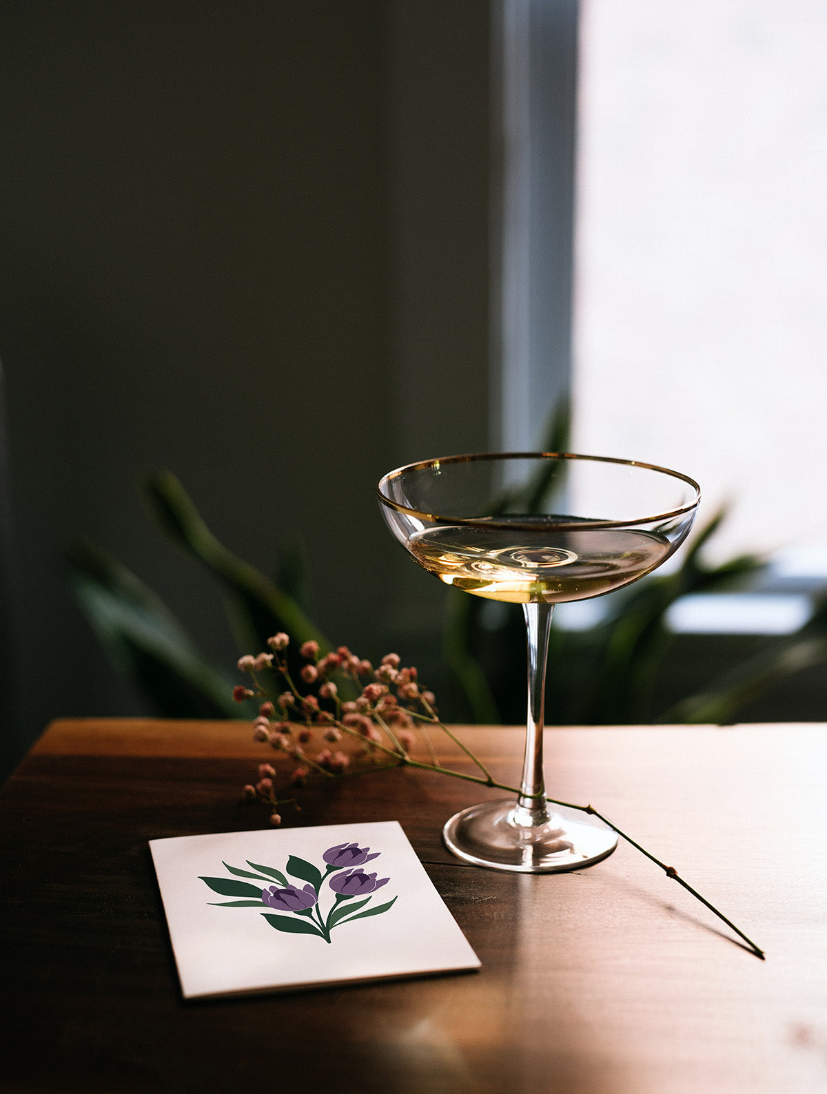 Hand-illustrated greeting card on a wooden table beside a coupe glass and dried flowers