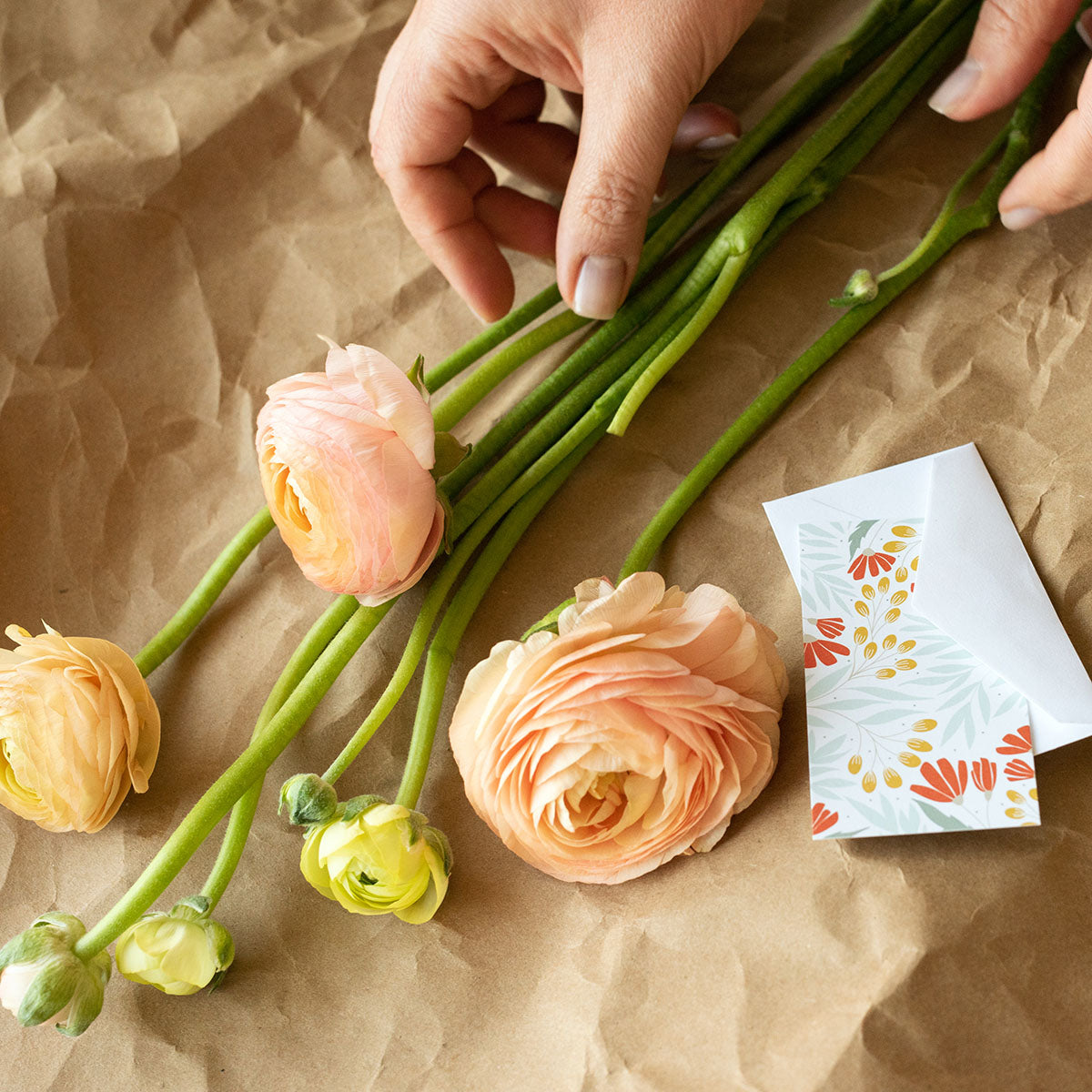 Hand arranging ranunculus flowers beside a Hazelmade mini note card on kraft paper