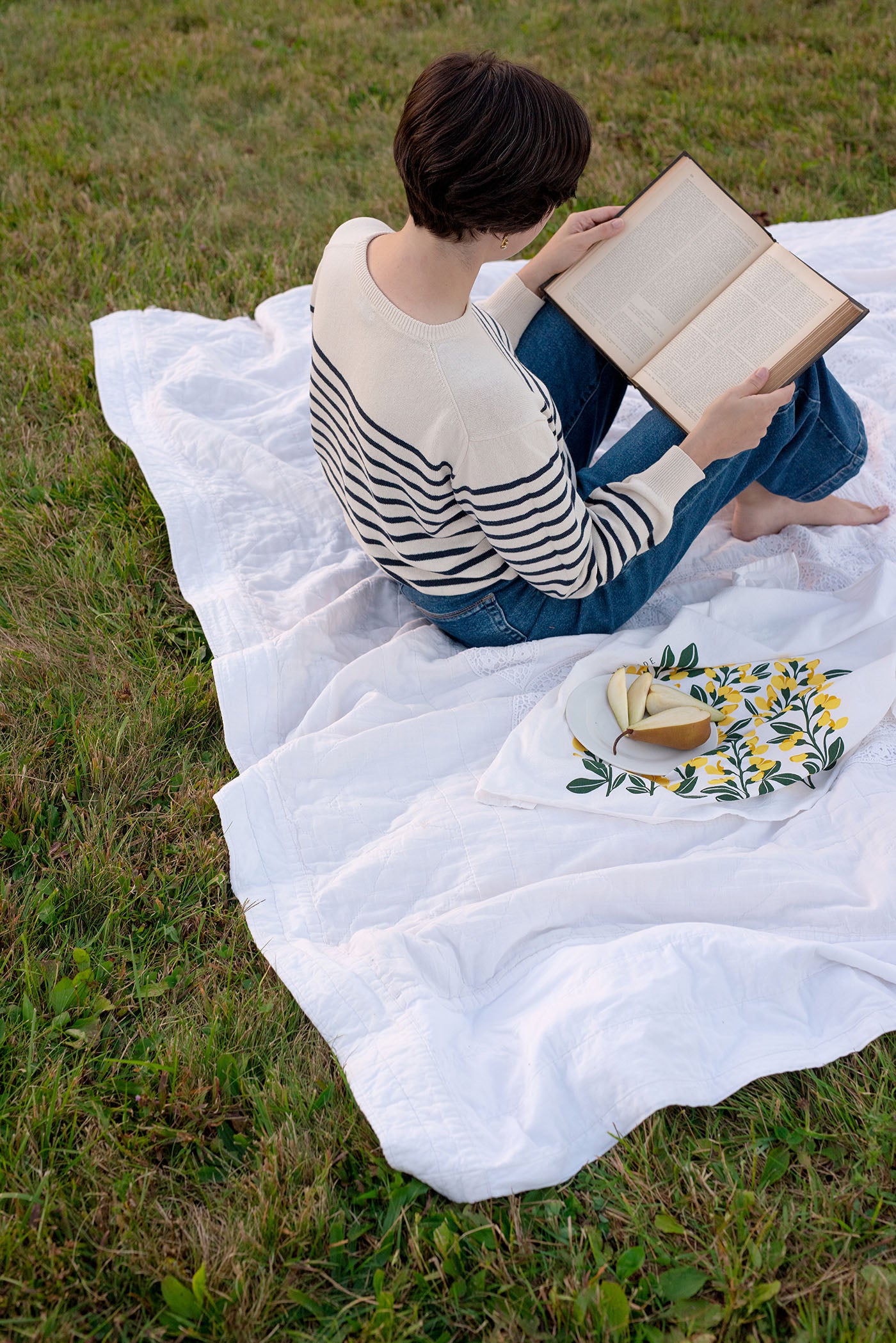 Person reading on a white blanket outdoors beside a hand-illustrated tea towel and sliced pear