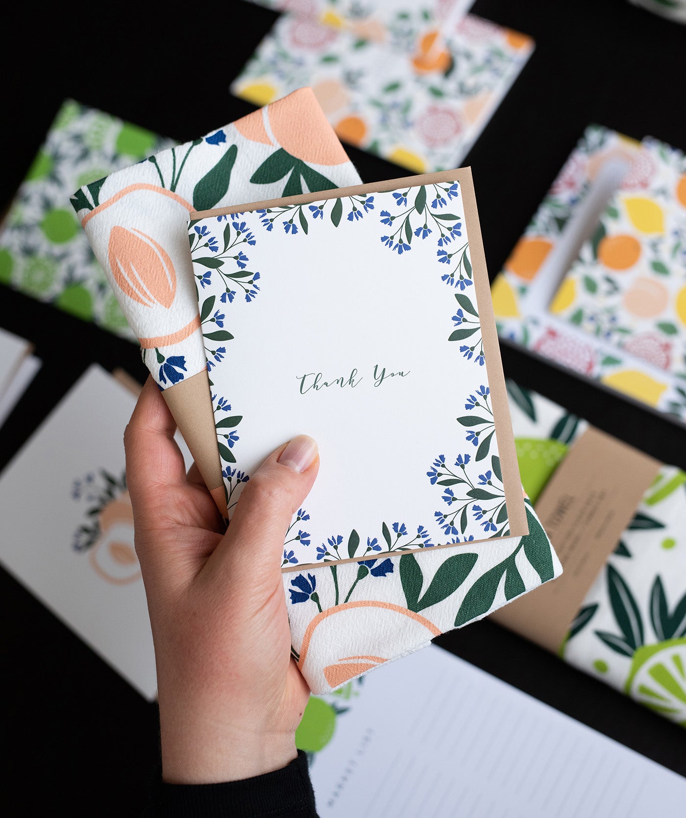 Hand holding a Hazelmade thank you card and coordinating tea towel above a collection of patterned paper goods