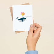 A hand holding the Happy Birthday Whale with Balloons greeting card and envelope showing the hand-illustrated whale and balloons design