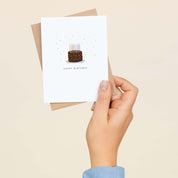 A hand holding a Happy Birthday Chocolate Cake greeting card and envelope showing the hand-illustrated chocolate cake with candles design