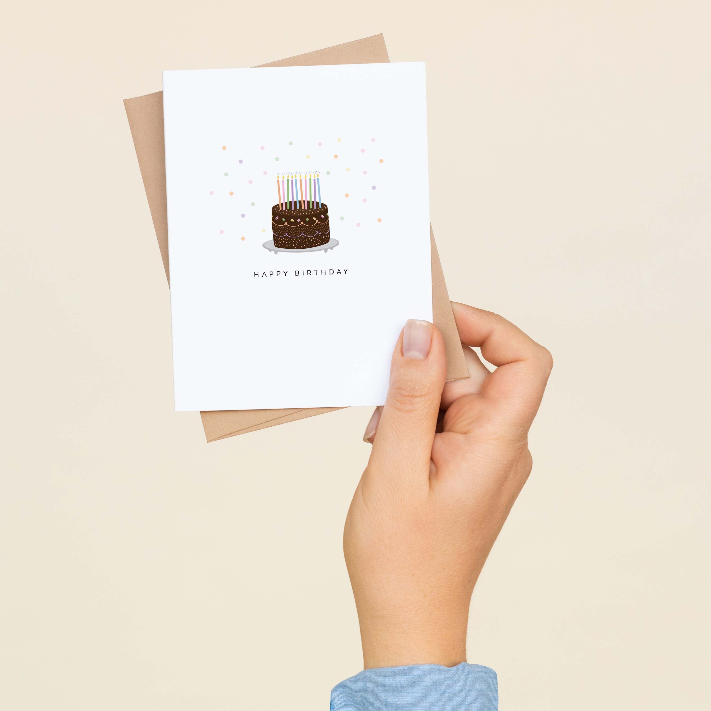A hand holding a Happy Birthday Chocolate Cake greeting card and envelope showing the hand-illustrated chocolate cake with candles design