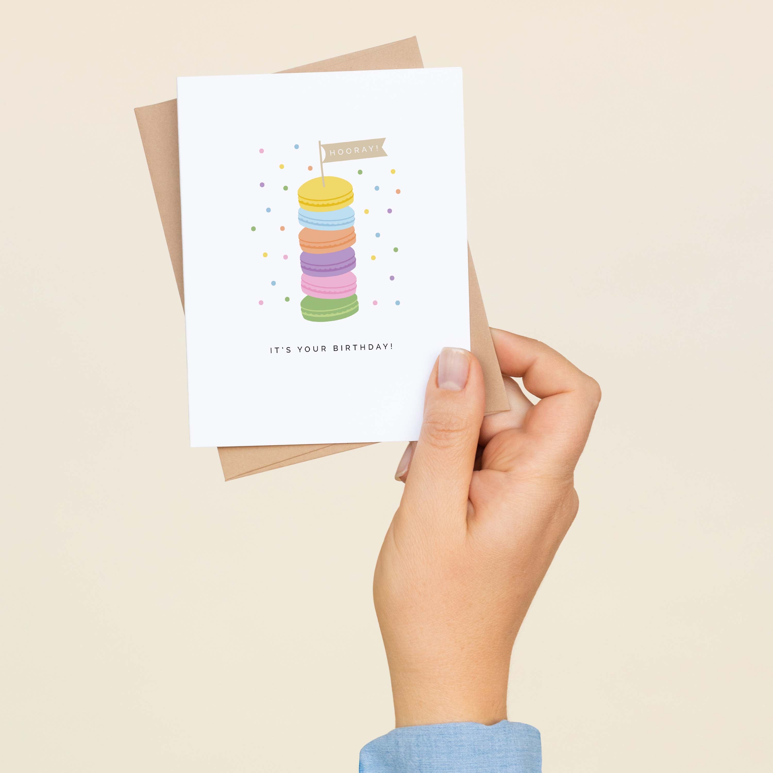 A hand holding the Hooray! It's Your Birthday! Macaron Stack greeting card and envelope showing the hand-illustrated colorful macaron stack design