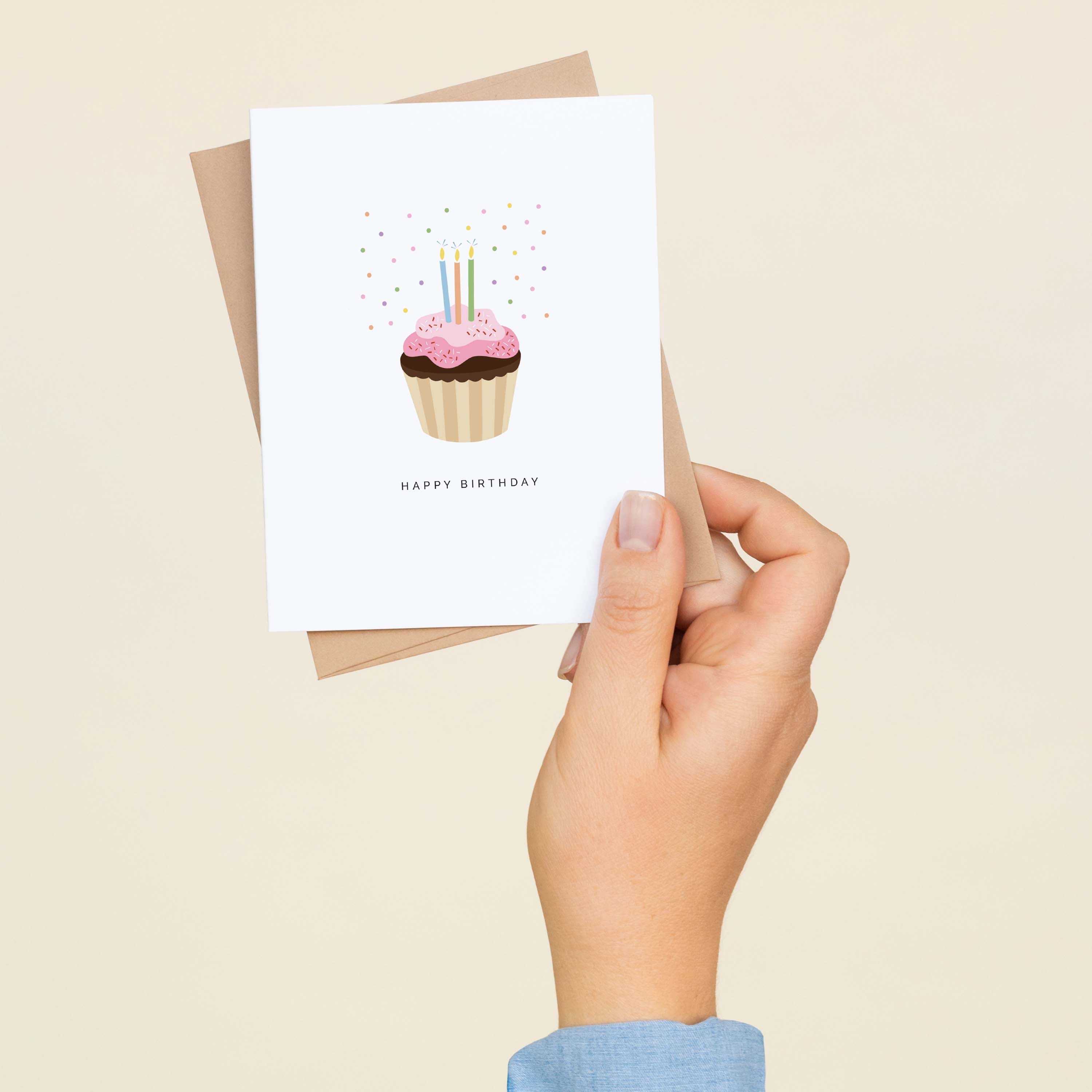 A hand holding a Happy Birthday Cupcake greeting card and envelope showing the hand-illustrated cupcake with candles design