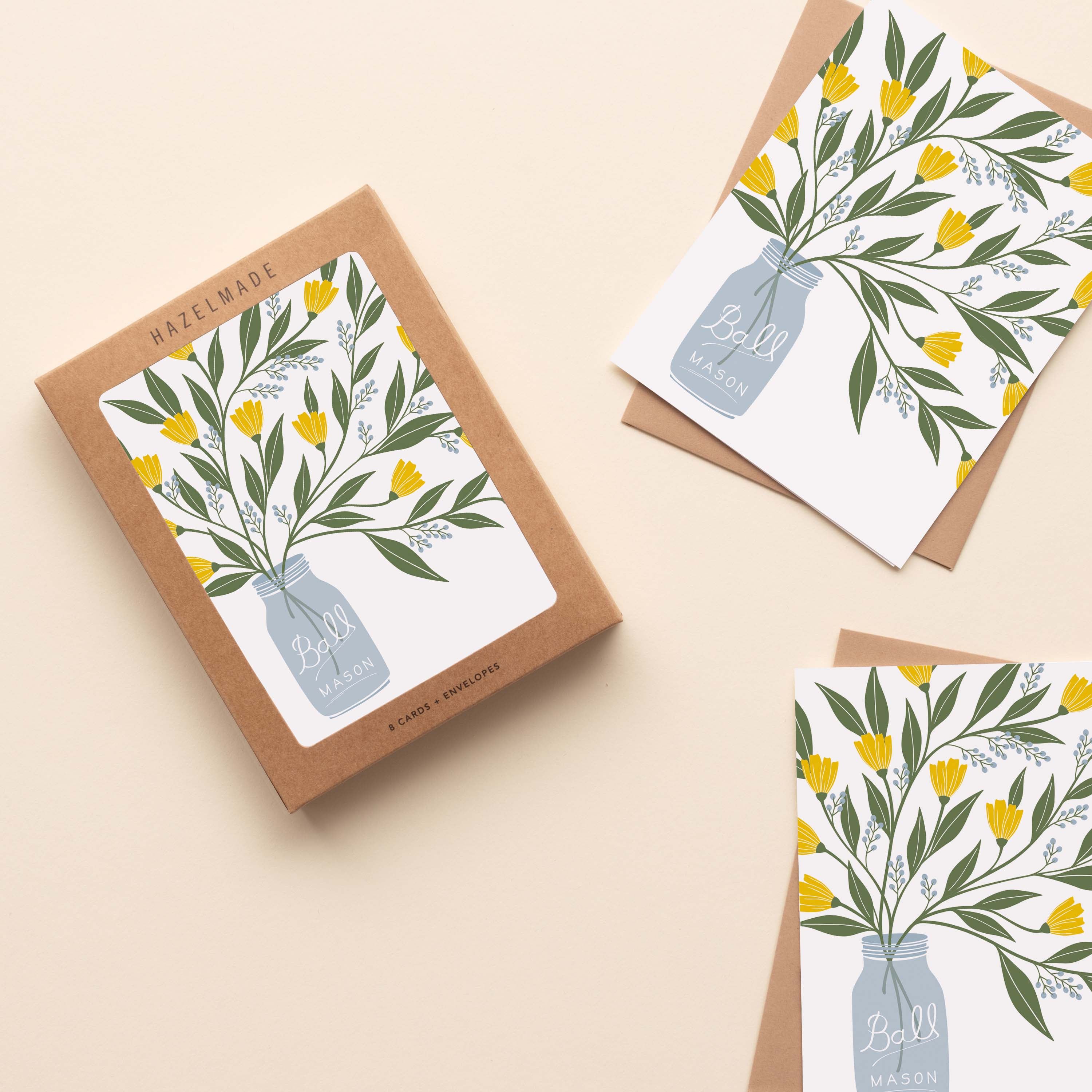A boxed set of Mason Jar Bouquet Greeting Cards sitting with two cards and their envelopes featuring a hand-illustrated mason jar filled with yellow flowers