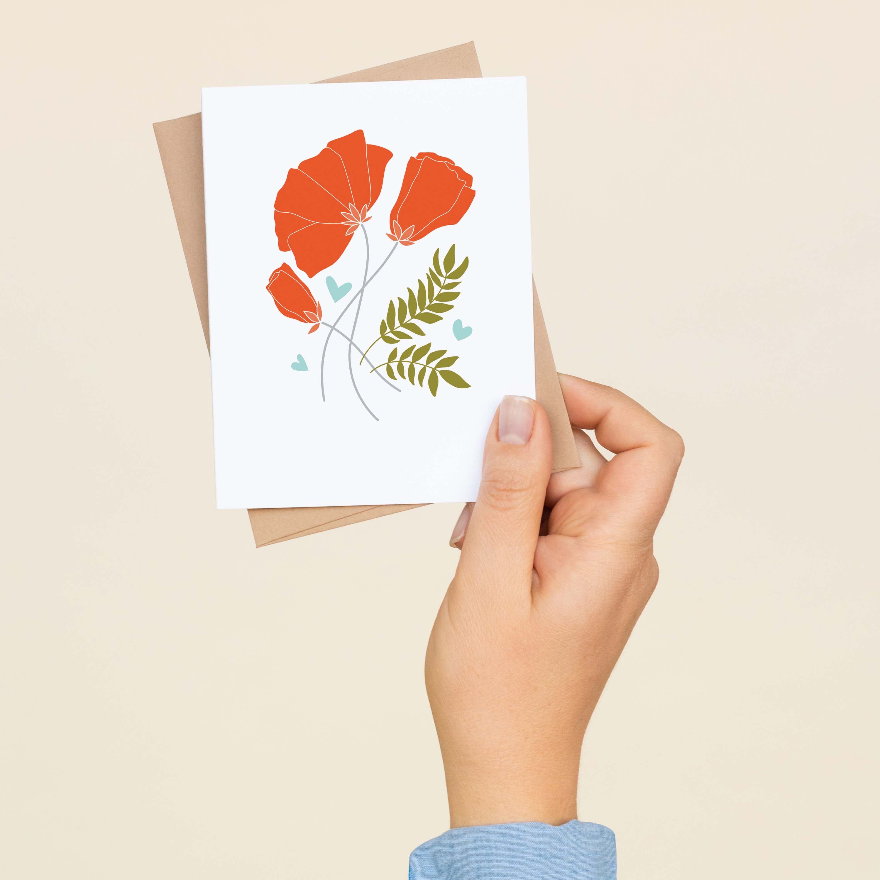 A hand holding a Poppies + Wheat greeting card and envelope showing the hand-illustrated poppies and wheat design