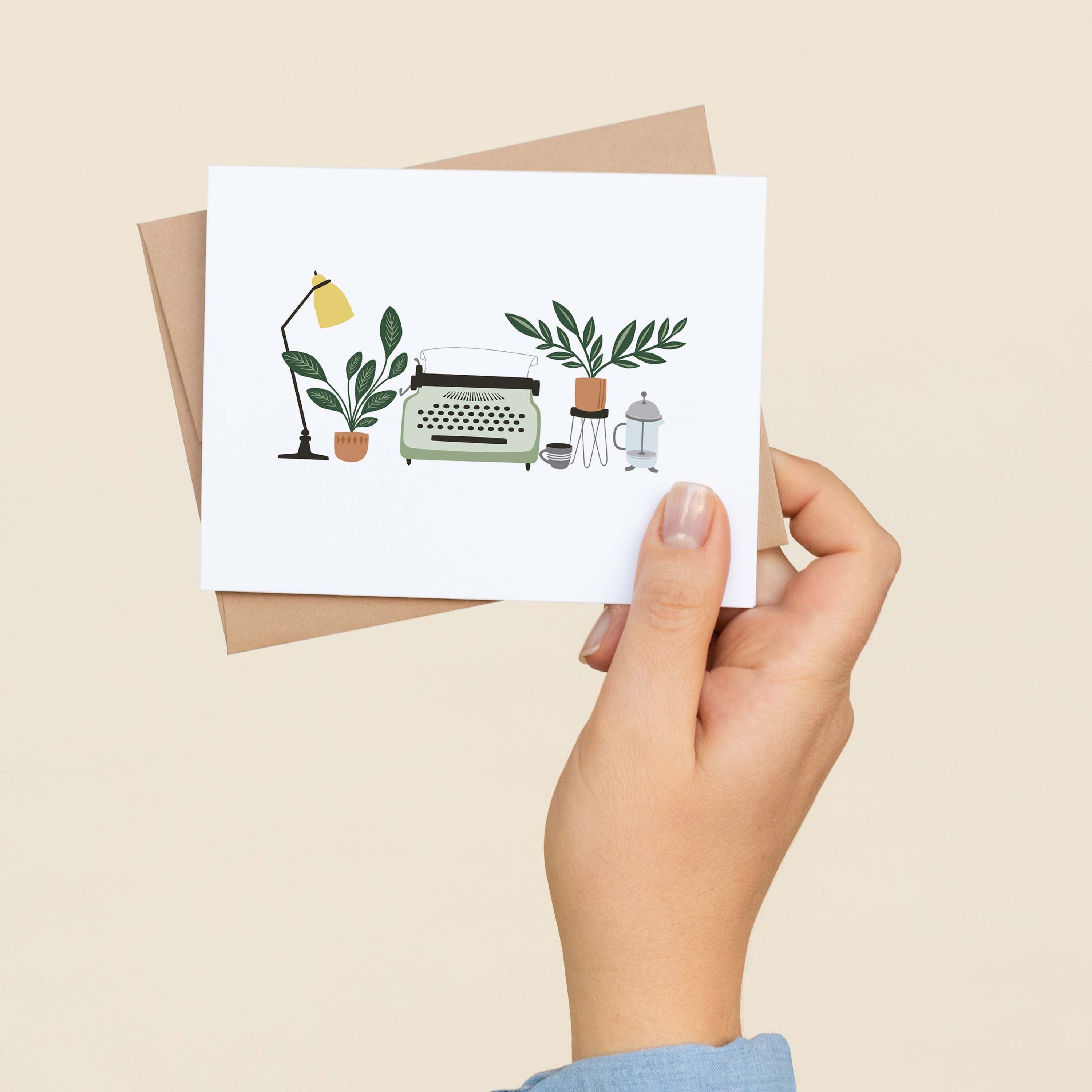 A hand holding a Typewriter & Desk greeting card and envelope showing the hand-illustrated typewriter and desk scene