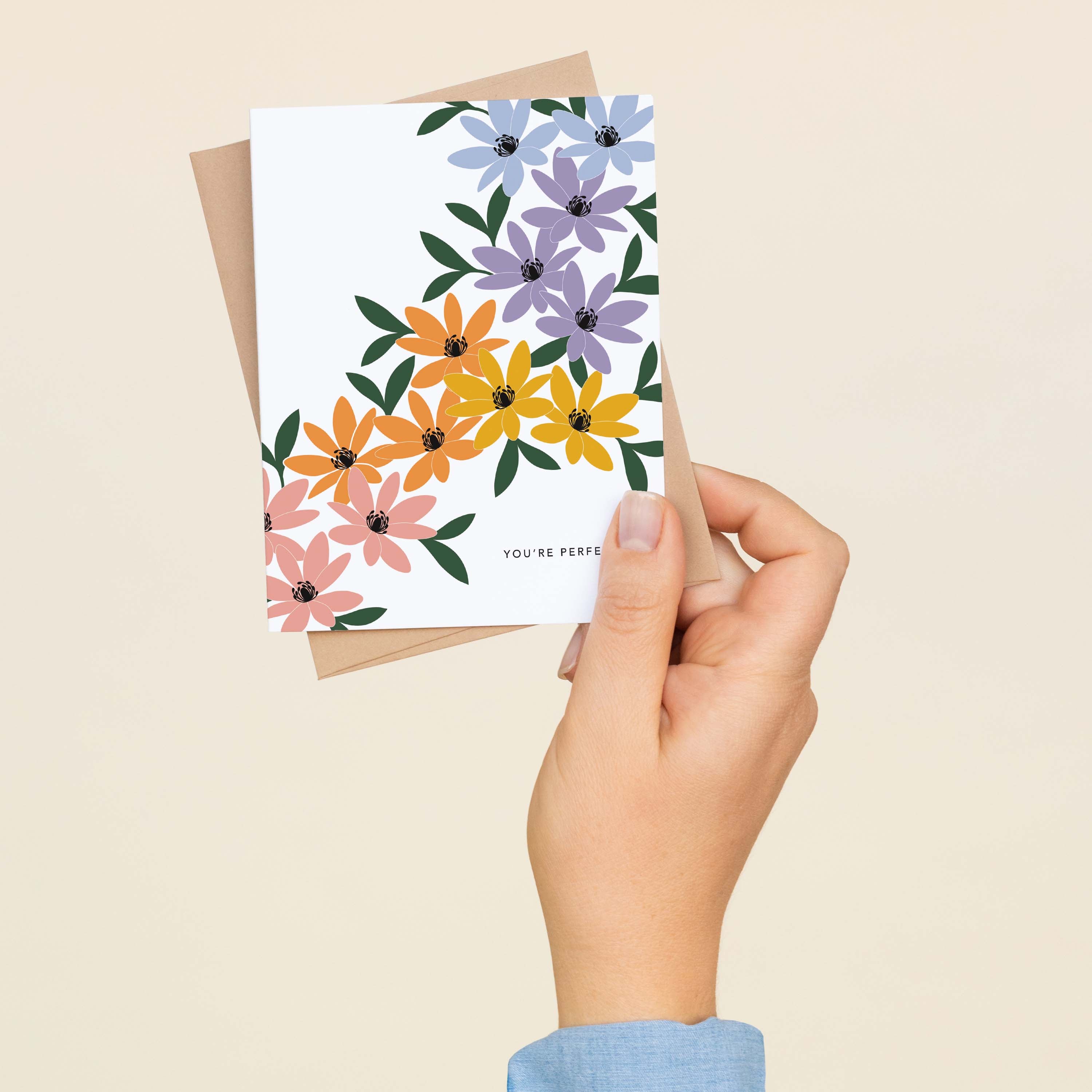 A hand holding the You're Perfect Rainbow Florals greeting card and envelope showing the hand-illustrated rainbow floral design