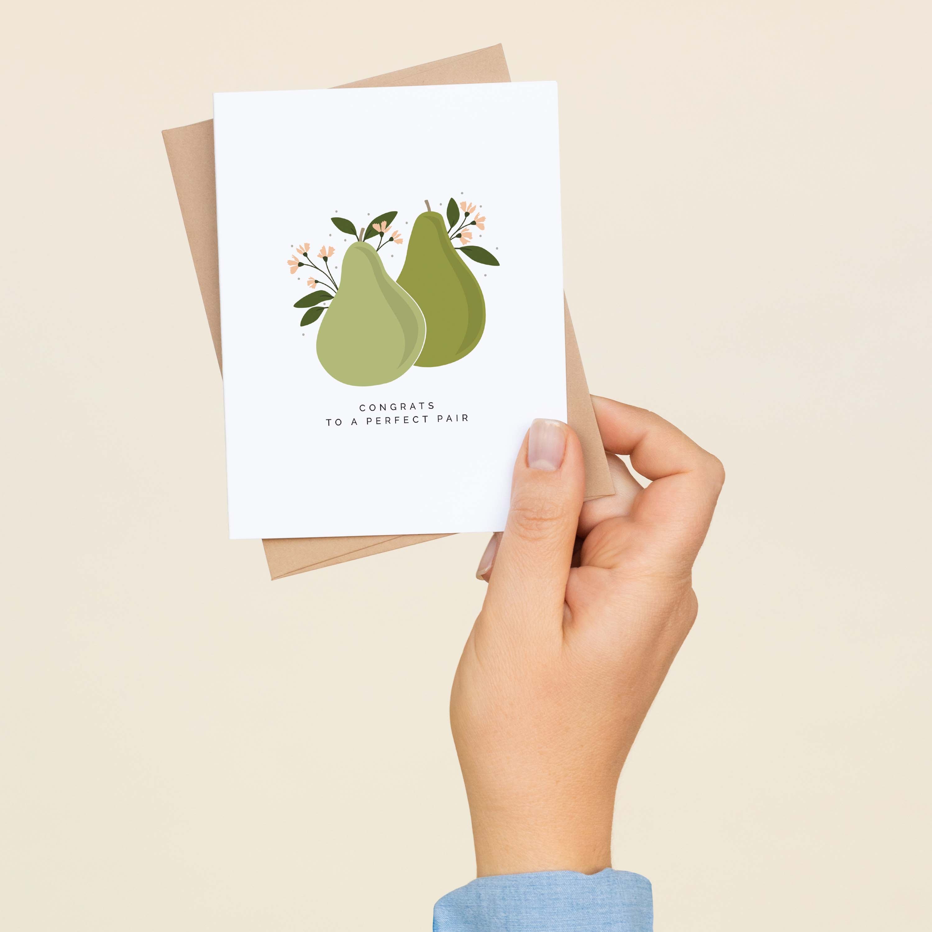 A hand holding the Congrats to a Perfect Pair greeting card and envelope showing the hand-illustrated pear design