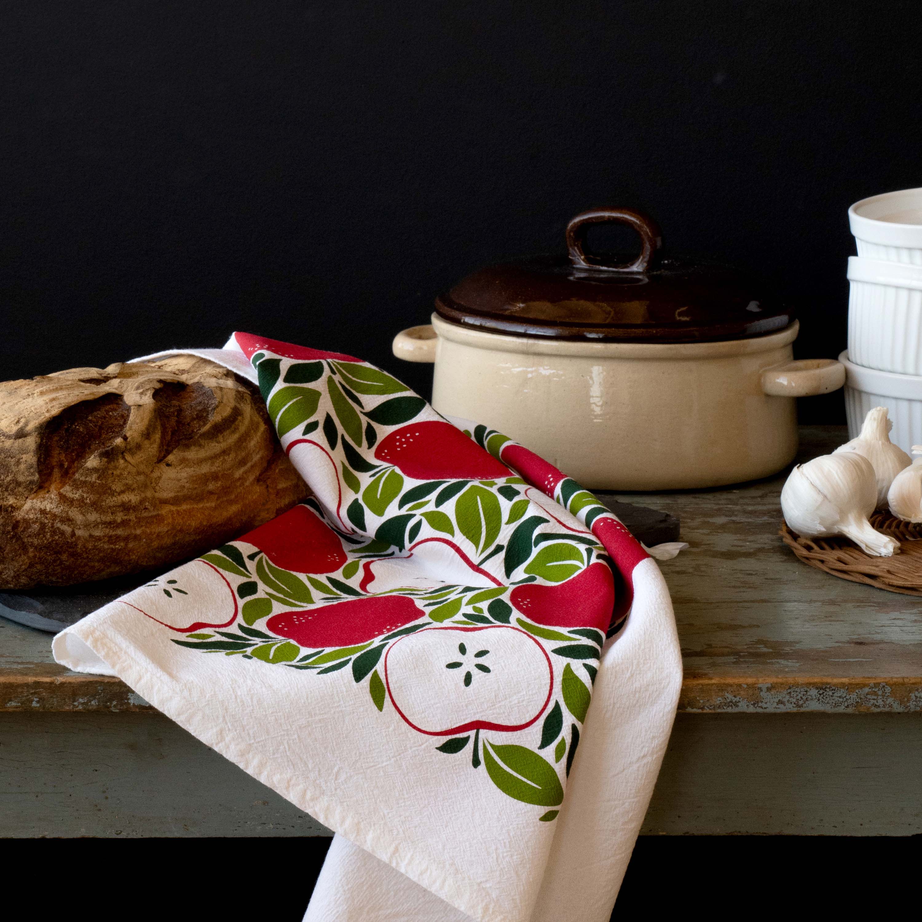 Harvest Apples Tea Towel draped over a table beside a Dutch oven, garlic, and bread