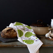 Artichokes + Olives Tea Towel styled beside bread, a casserole pot, and garlic on a dark table