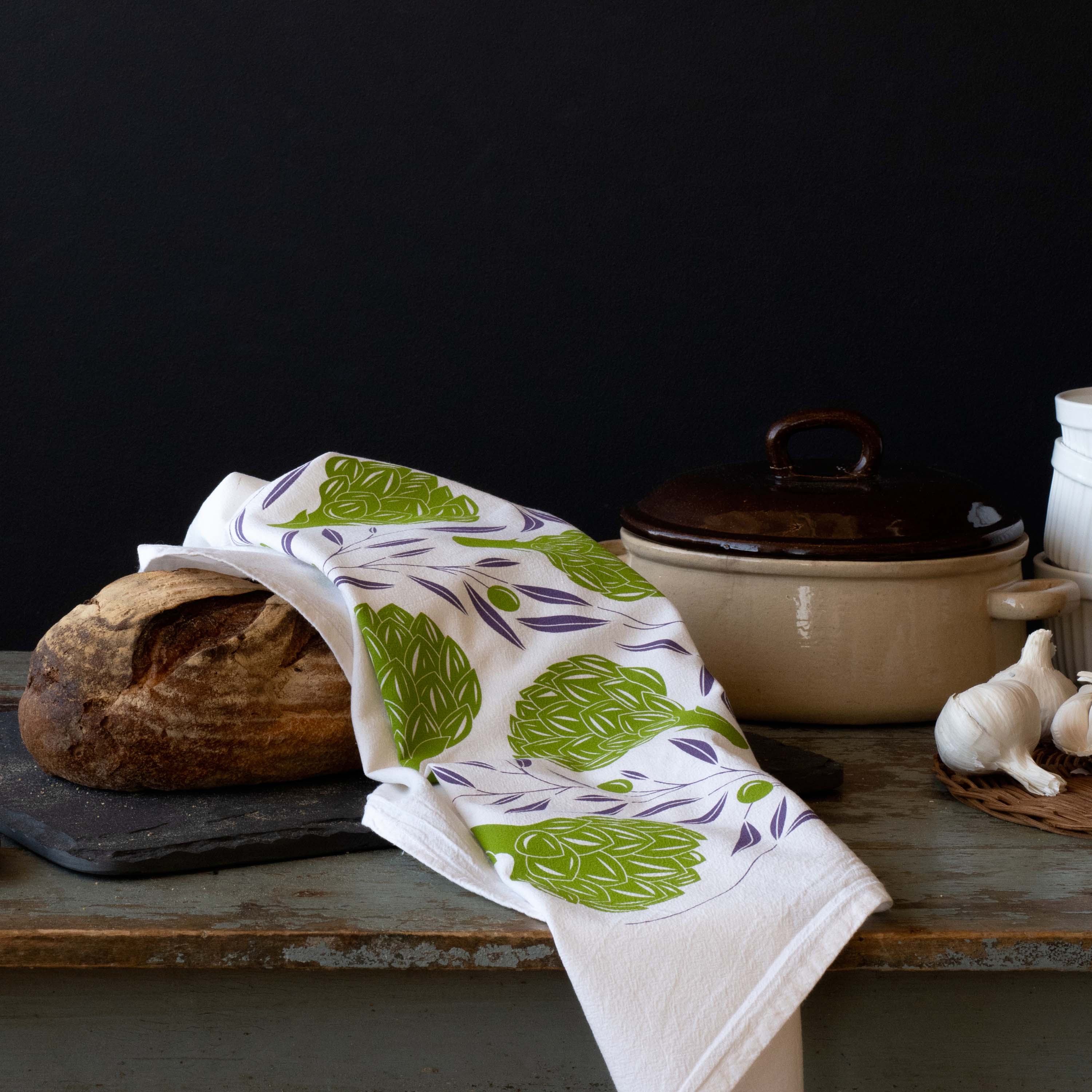 Artichokes + Olives Tea Towel styled beside bread, a casserole pot, and garlic on a dark table