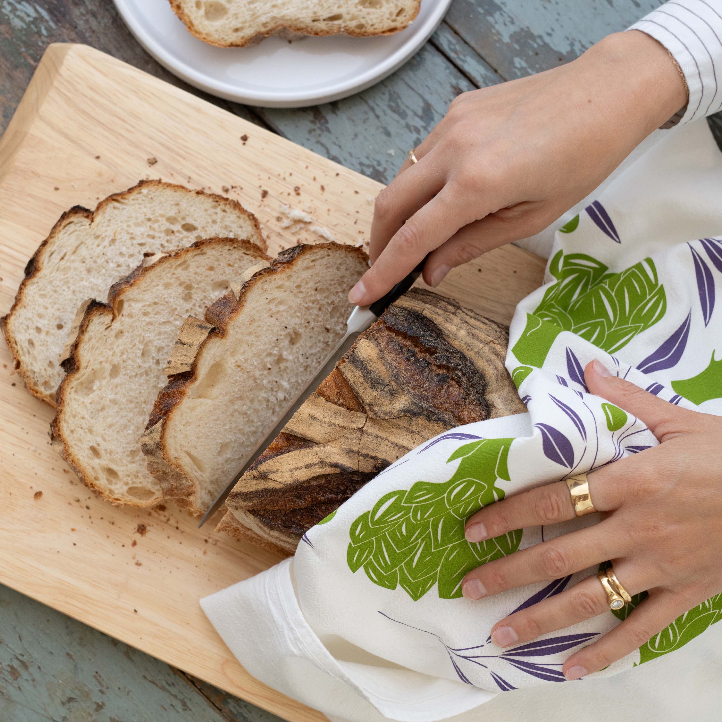 Artichokes + Olives Tea Towel wrapped around a loaf of sliced bread while a person cuts it on a wooden board