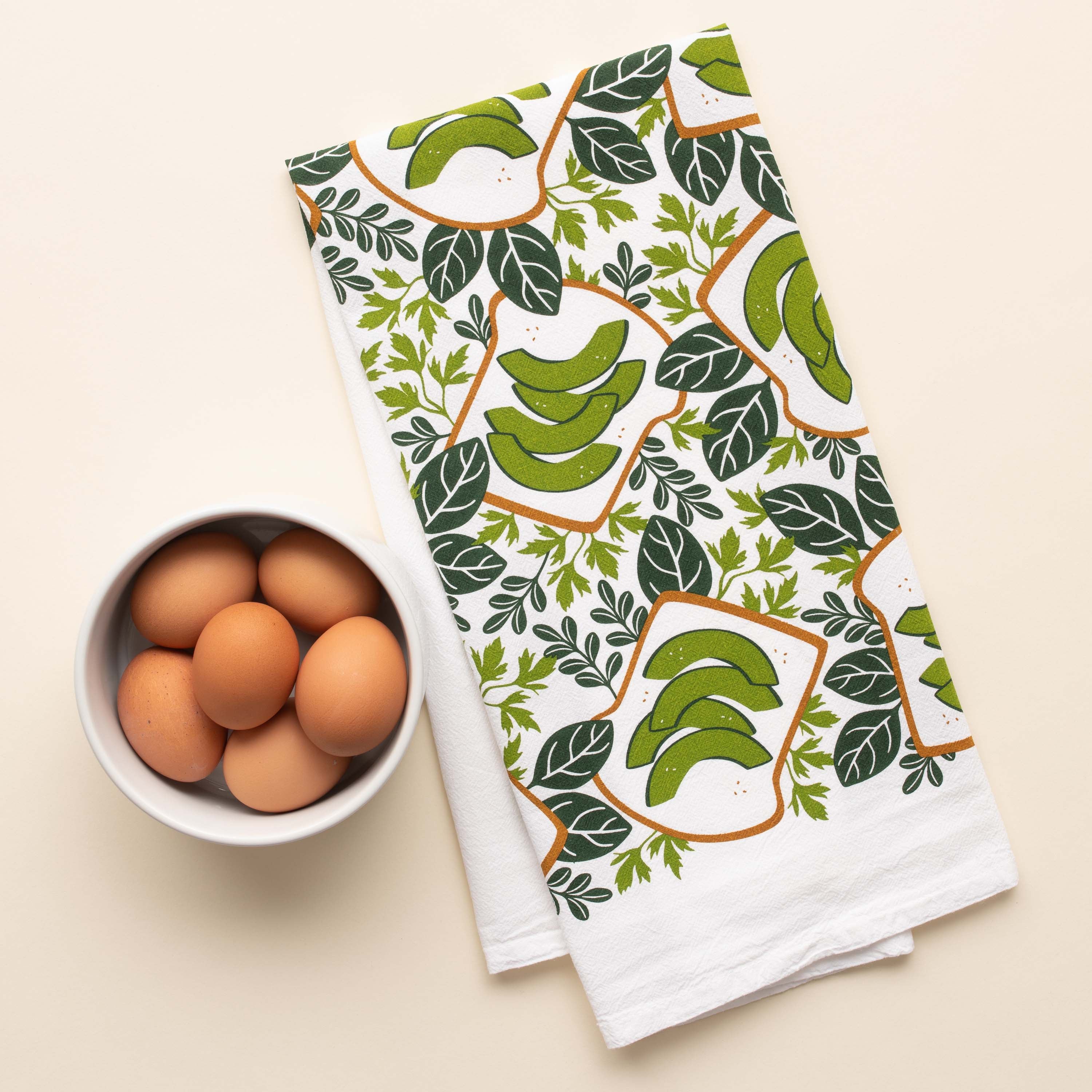 Avocado Toast Tea Towel with hand-illustrated avocado toast design beside a bowl of brown eggs