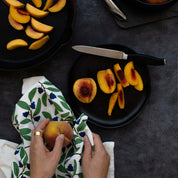 Blueberry Bushel Tea Towel styled beside sliced peaches on a dark tabletop