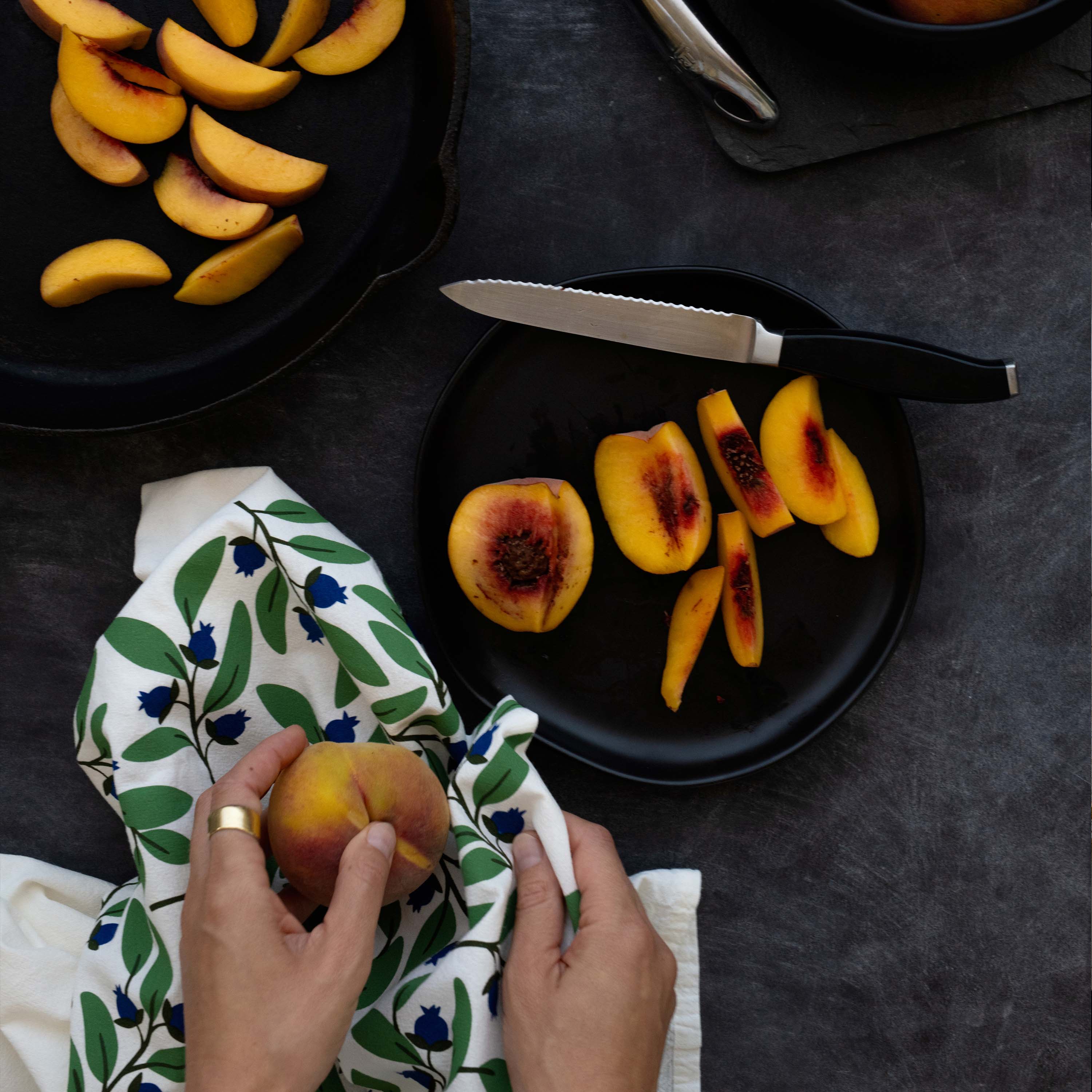 Blueberry Bushel Tea Towel styled beside sliced peaches on a dark tabletop