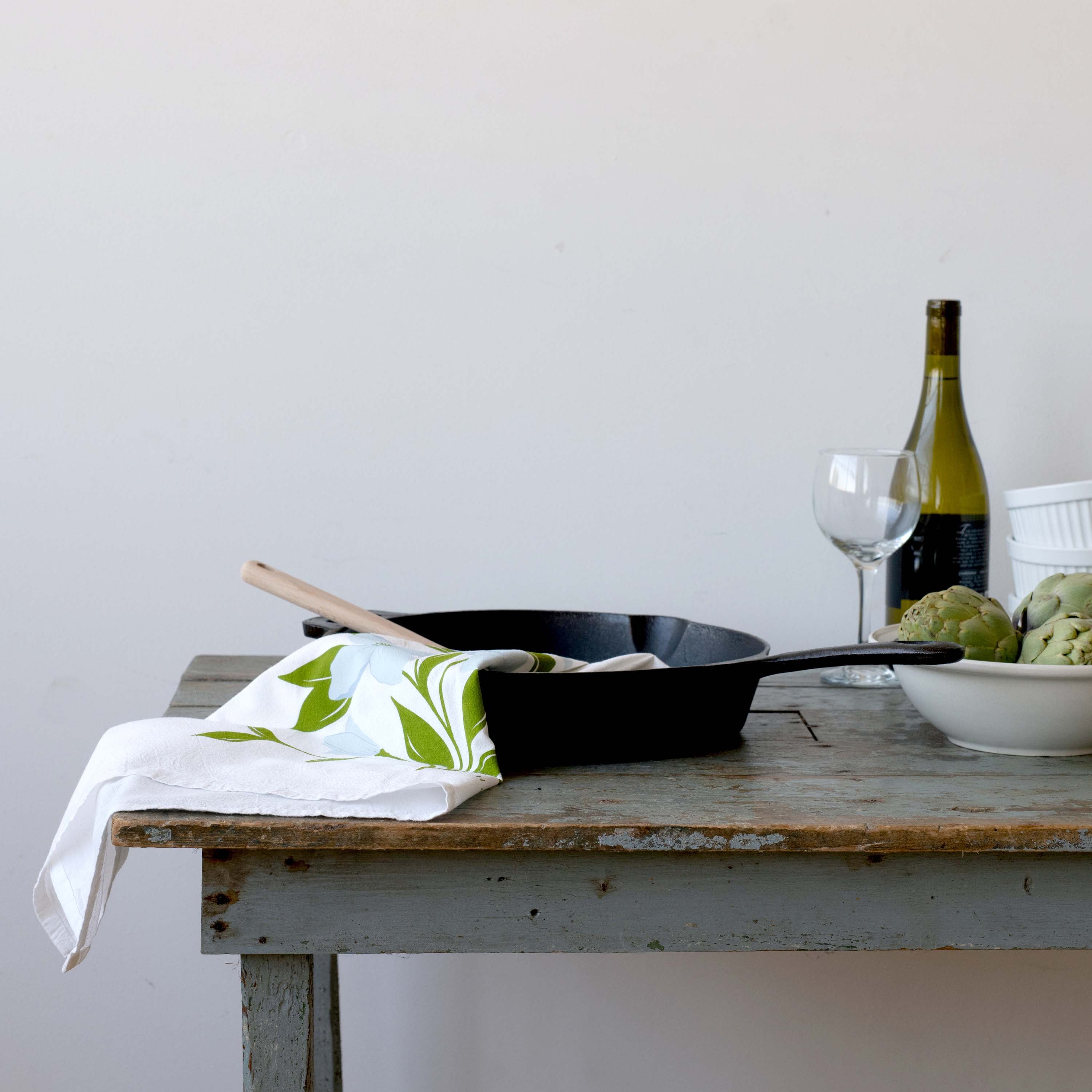 Dogwood Tea Towel styled on a rustic table with a skillet, a bottle of wine and artichokes