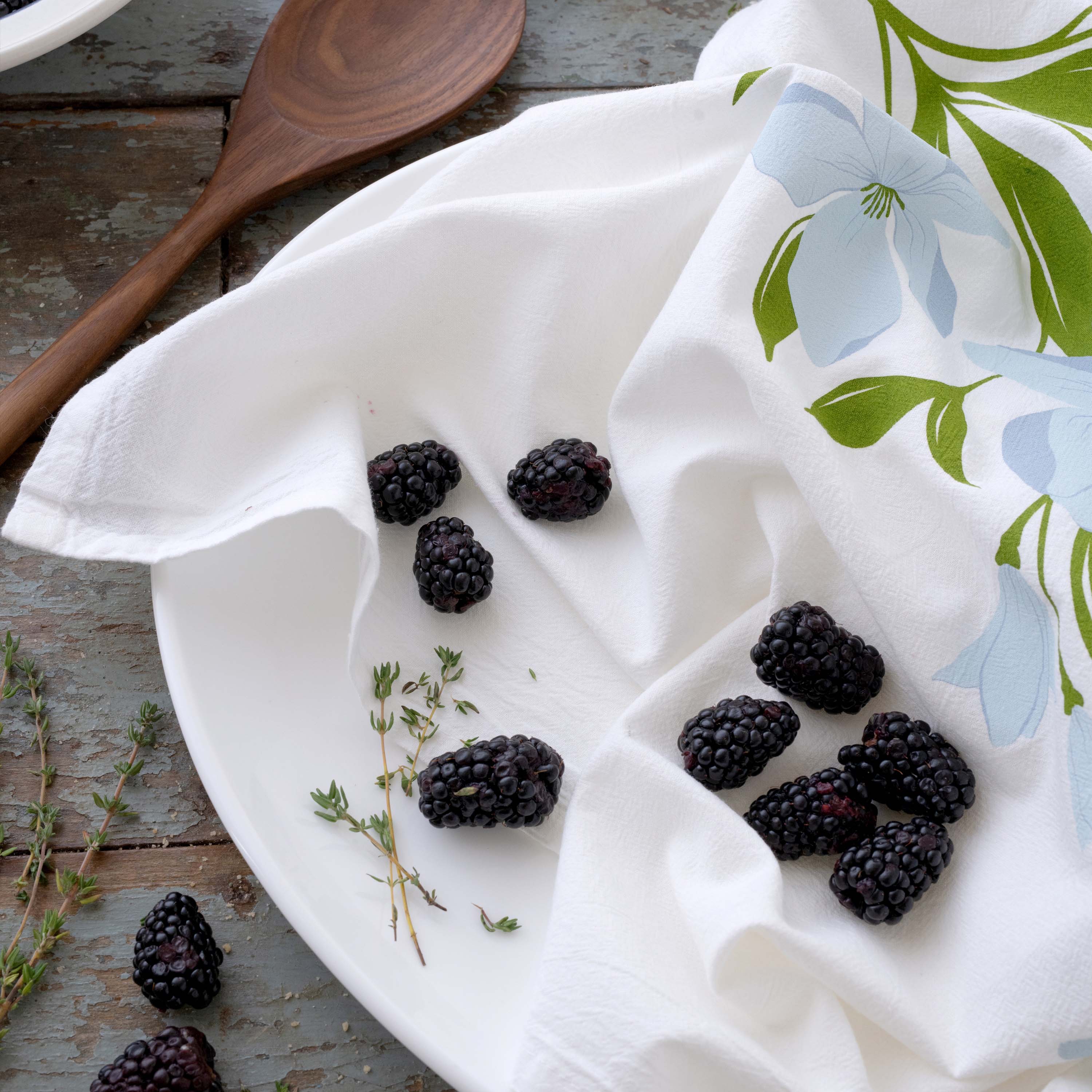 Dogwood Tea Towel styled with blackberries and a wooden spoon on a wooden tabletop