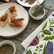 Purple Farmers Market Tea Towel styled beside a plate of crostini and a bowl of herbs and pepper