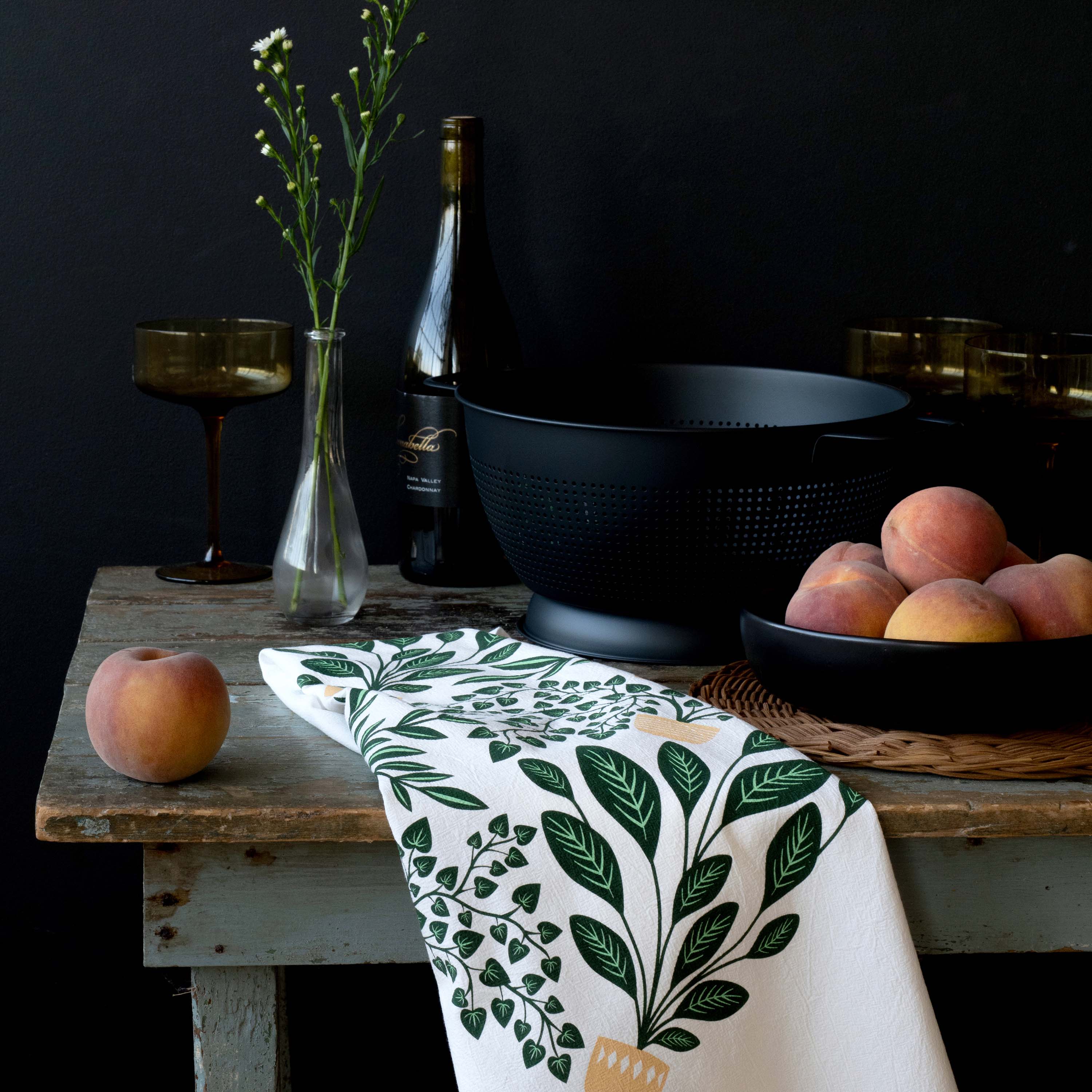 House Plants Tea Towel styled on a rustic table with peaches, glassware, and dark cookware