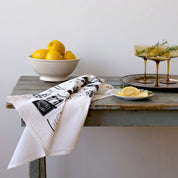 Italian Market Tea Towel draped over a wooden table beside lemons and drinks
