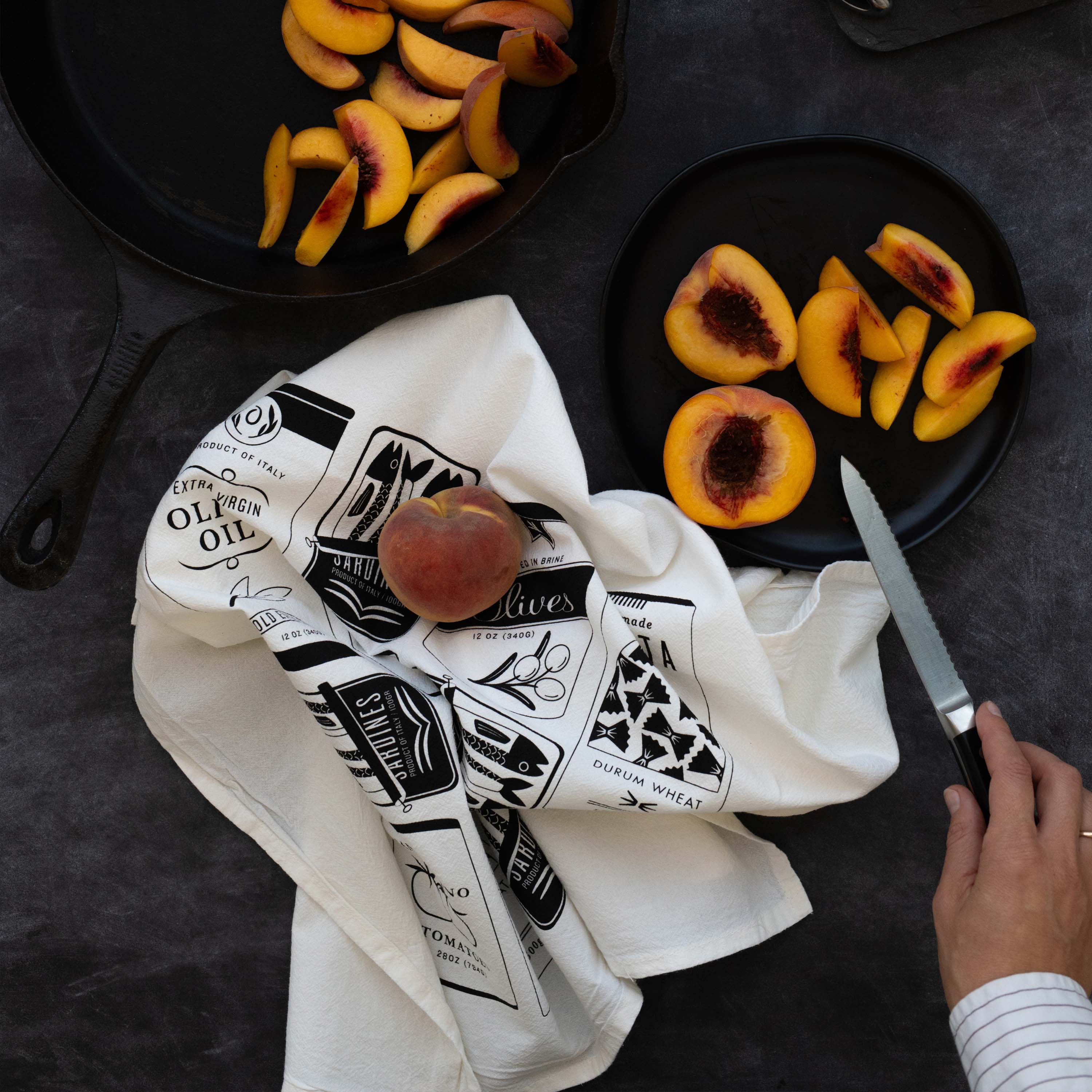 Italian Market Tea Towel styled beside sliced peaches while someone slices peaches on a dark tabletop