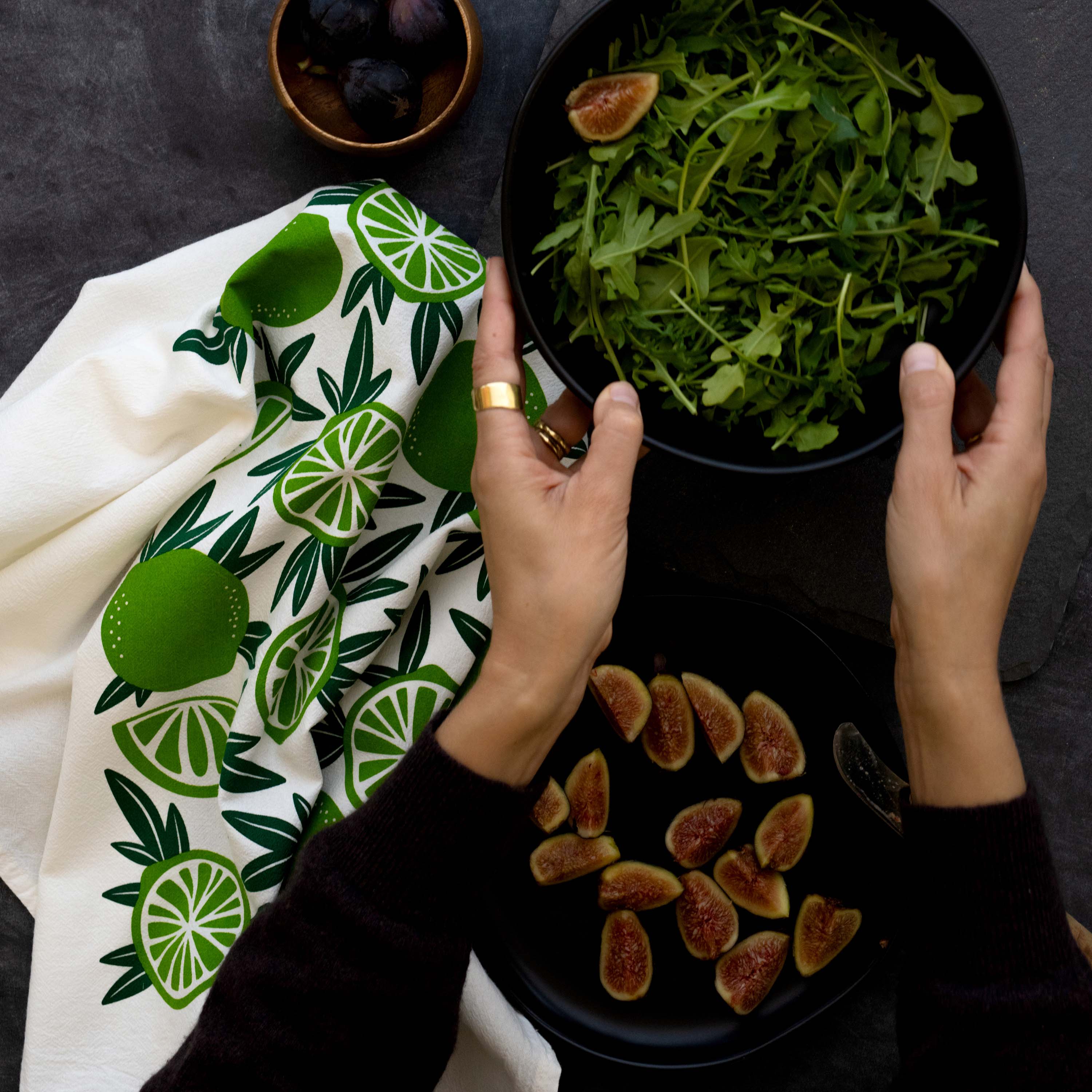 Limes Tea Towel on a dark table next to a plat of figs and a bowl of greens held in both hands