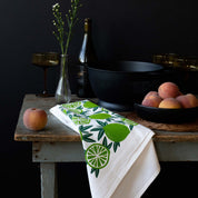 Limes Tea Towel draped over a table beside a wine bottle, a colander and bowl of peaches