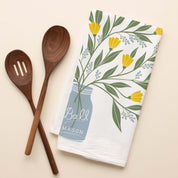 Mason Jar Bouquet Tea Towel with hand-illustrated mason jar and yellow floral bouquet beside wooden kitchen utensils