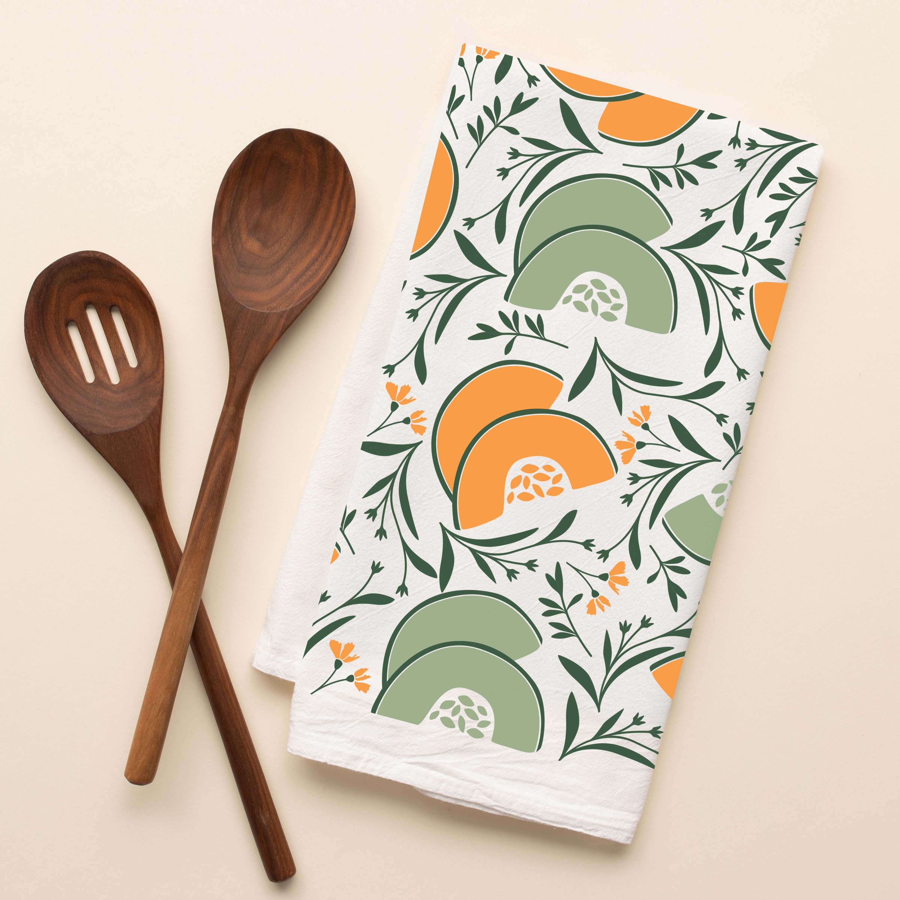 Cantaloupe + Honeydew Tea Towel with hand-illustrated melon design beside wooden kitchen utensils