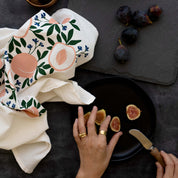 Peaches Tea Towel styled on a dark table with figs and a person arranging cutting figs