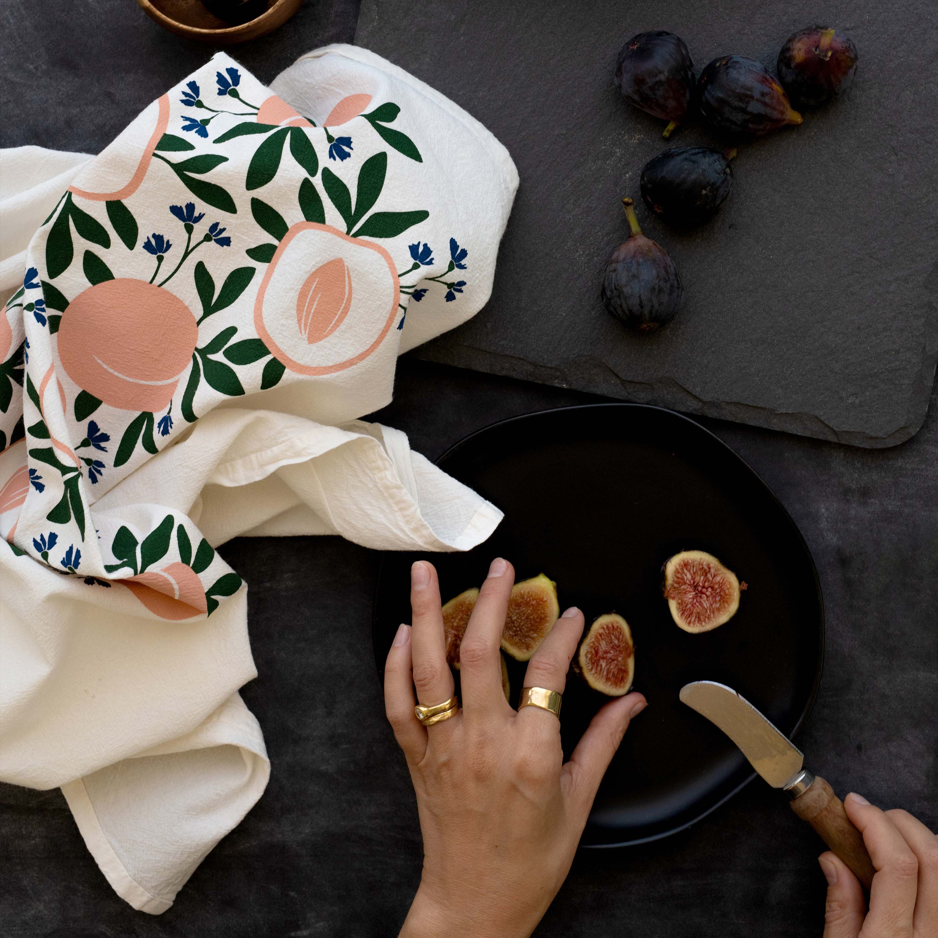 Peaches Tea Towel styled on a dark table with figs and a person arranging cutting figs