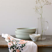 Peaches Tea Towel draped beside stacked bowls and a glass vase on a wooden table