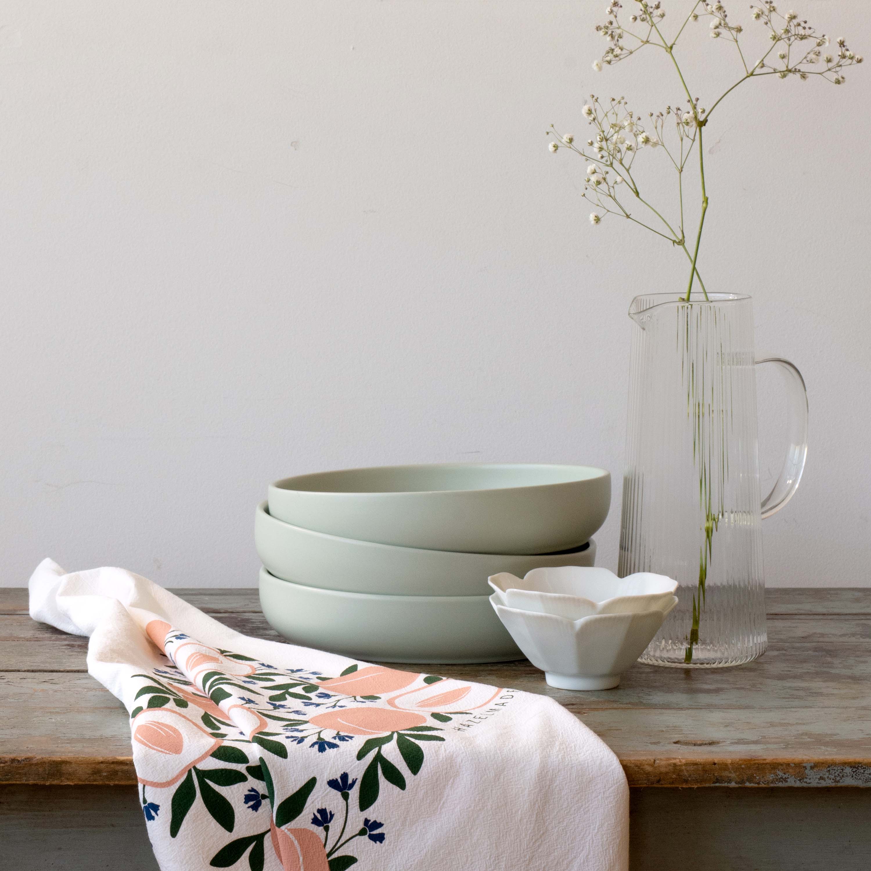 Peaches Tea Towel draped beside stacked bowls and a glass vase on a wooden table