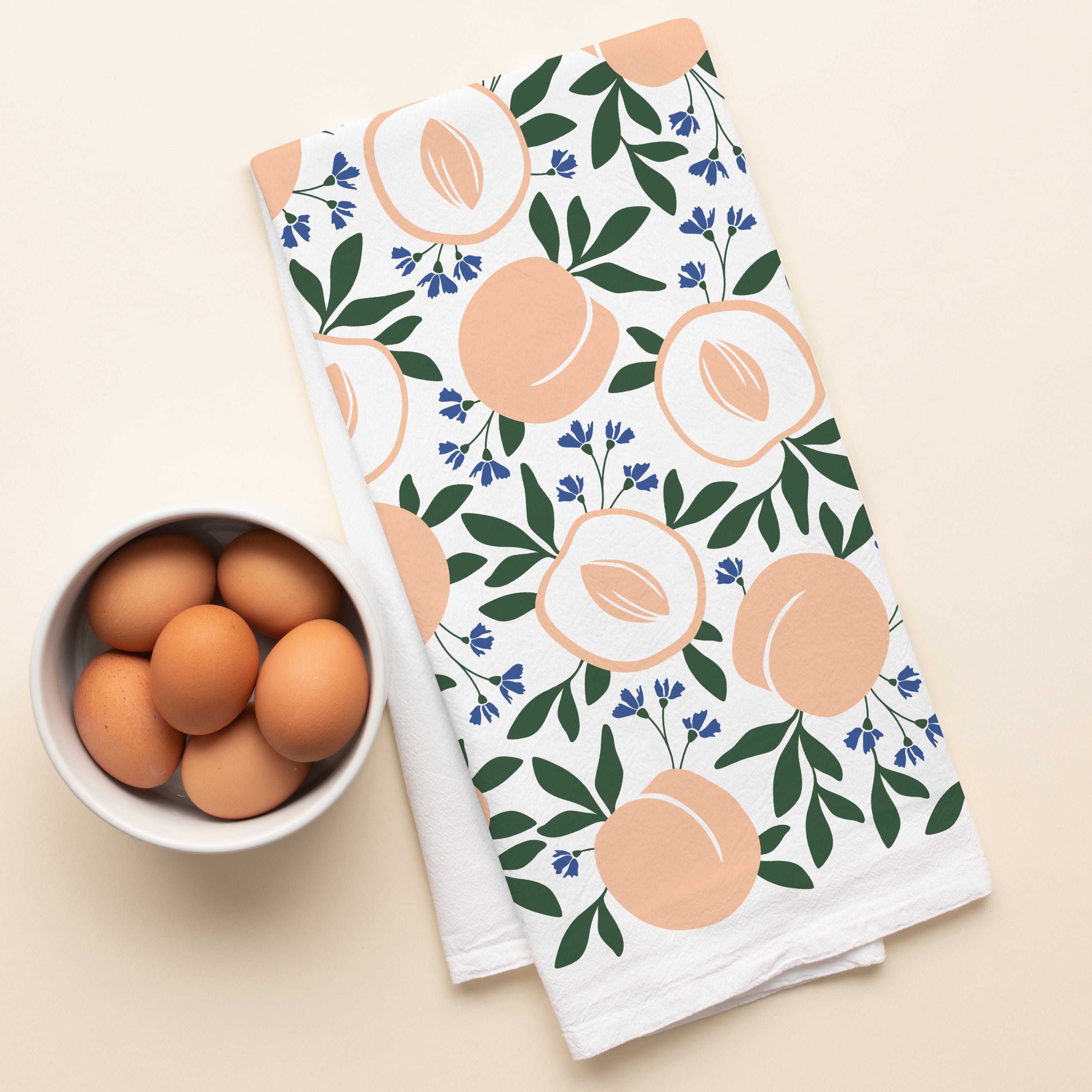 Peaches Tea Towel laid beside a bowl of brown eggs on a light surface